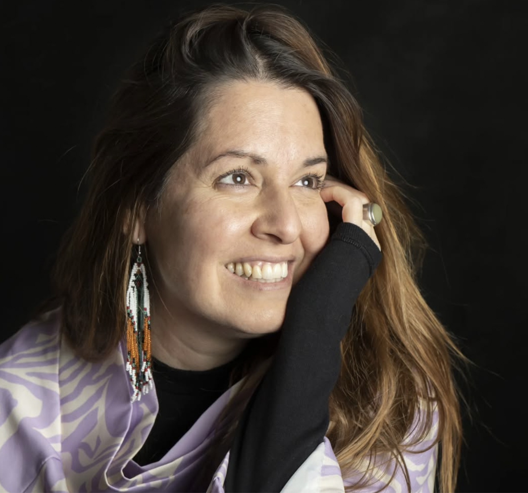 Mujer sonriendo, con cabello largo y ondulado, usando aretes de cuentas de colores y ropa con estampado en tonos morados y lavanda, con fondo negro.