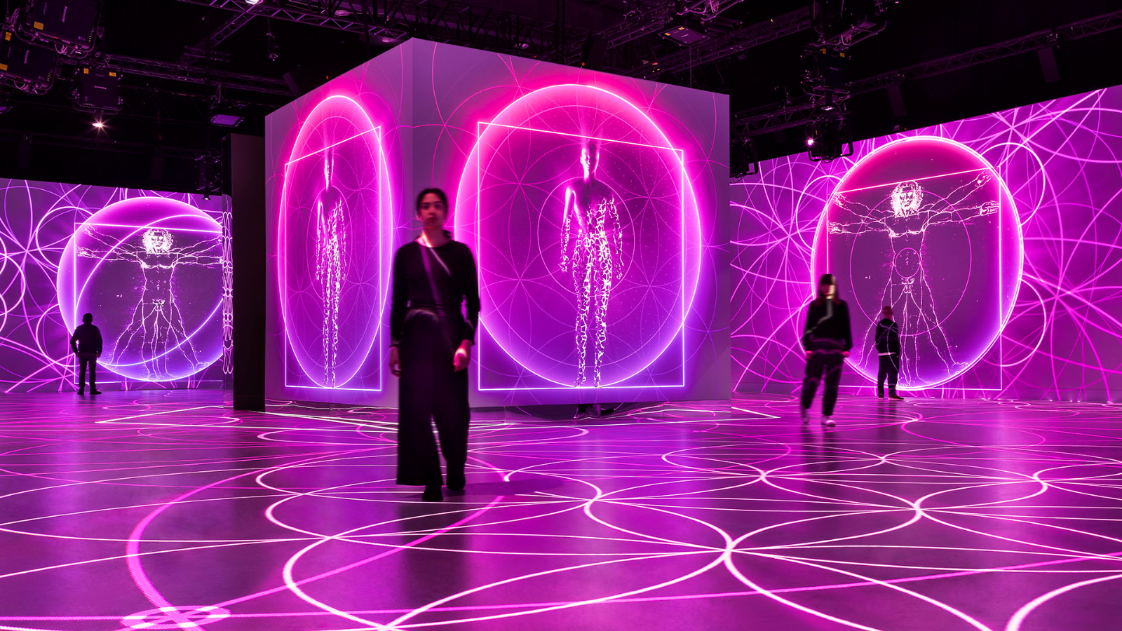 An interactive digital art installation featuring neon purple outlines of the human body, with glowing geometric patterns projections on the floor and walls, and people observing the display.