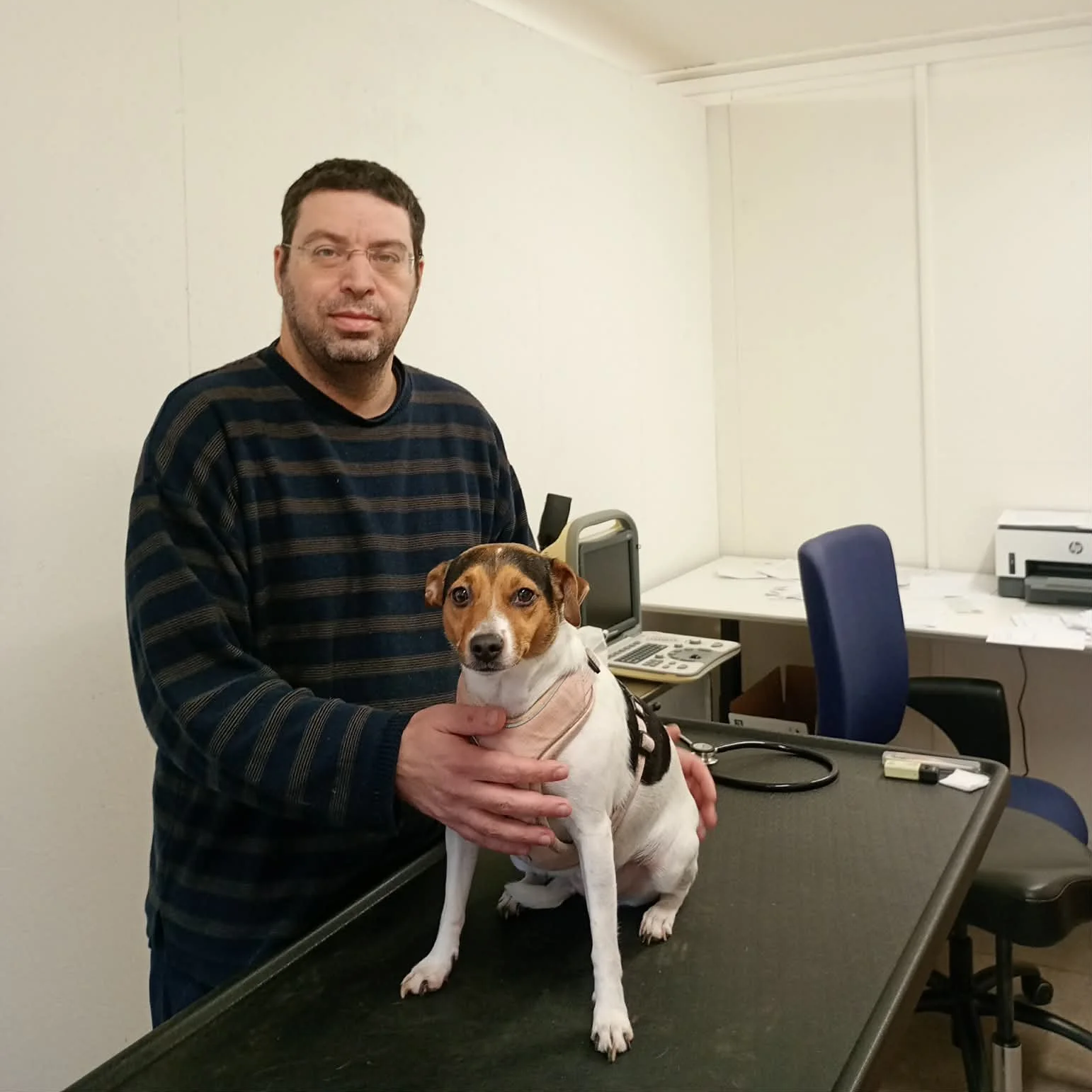A man in a striped sweater holding a small dog on an examination table in an office or clinic