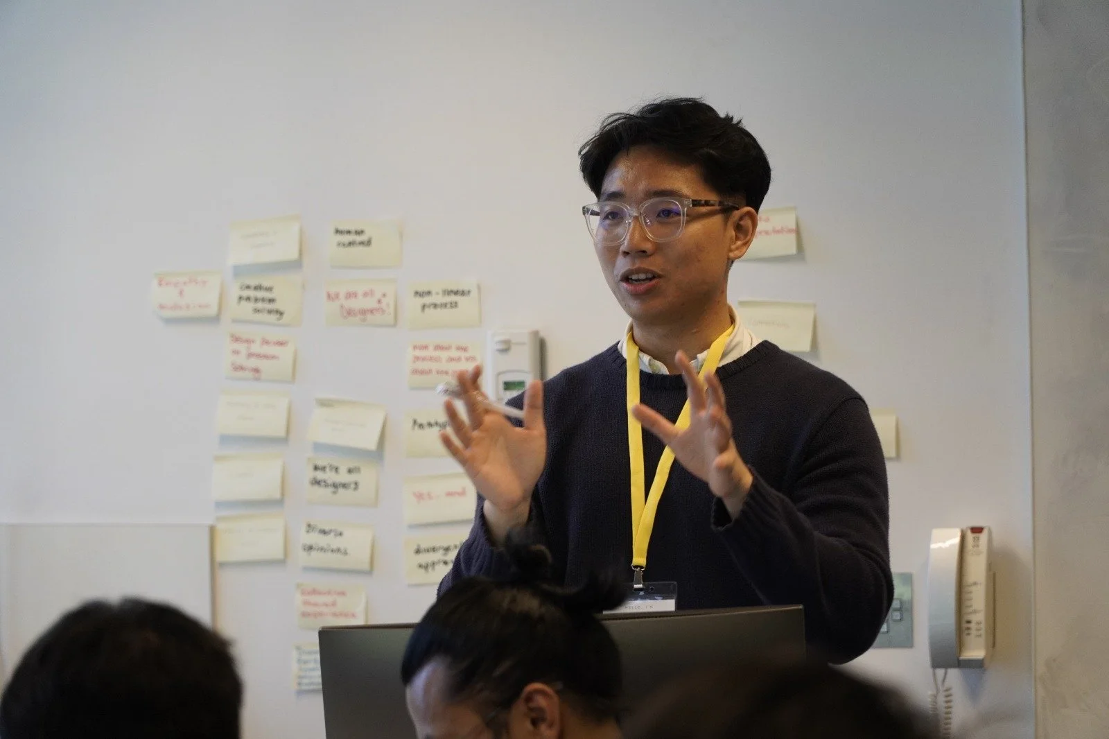 Young man with glasses speaking in front of a whiteboard covered with sticky notes during a presentation or workshop.