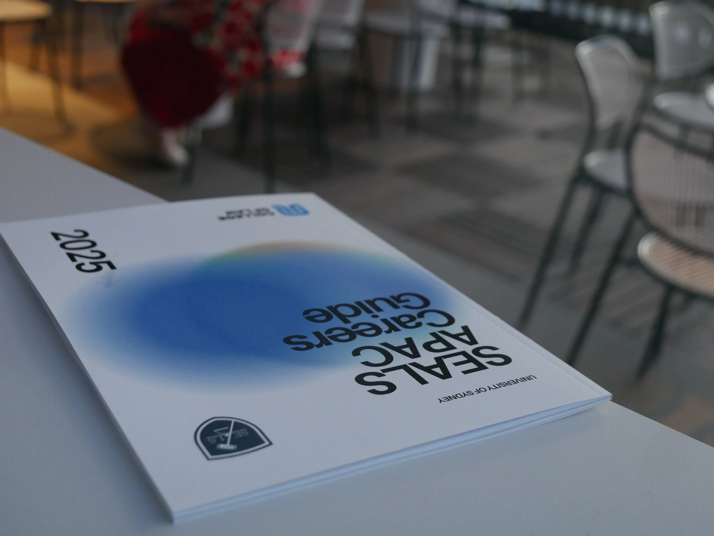 Book titled 'Seallas Arcada Calé Serve Guide' on a white table at a restaurant or café, with empty chairs and tables in the background.