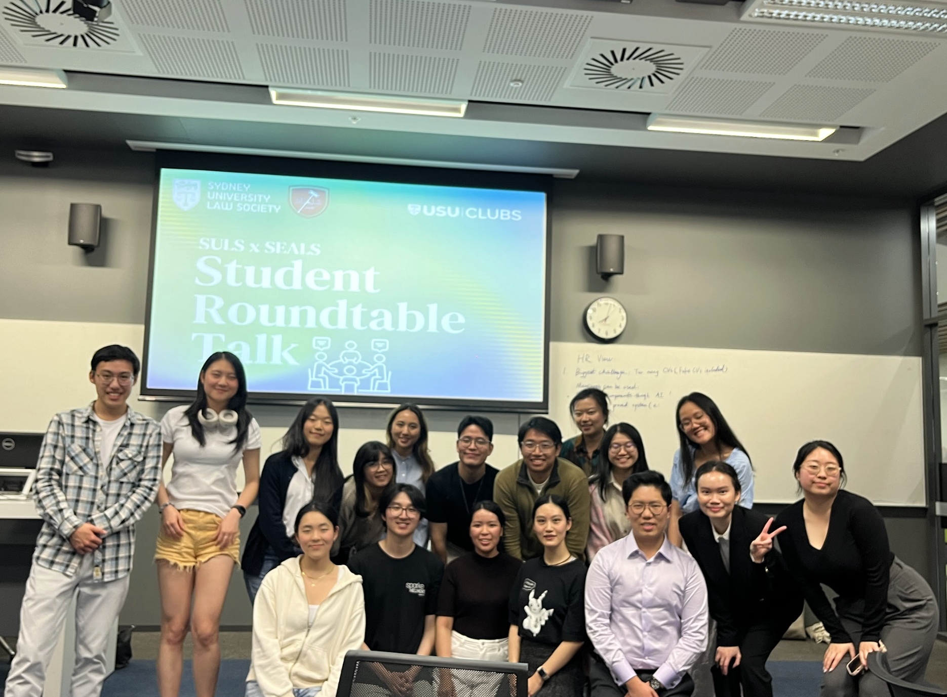 Group of diverse students posing for a photo in a classroom with a large screen displaying a presentation titled 'Student Roundtable Talk' by Sydney University Law Society and USU Clubs.