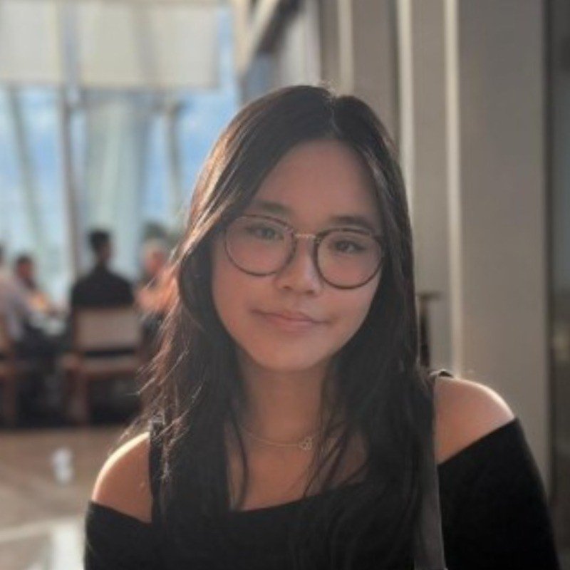 A young woman with dark hair and glasses sitting in a bright indoor space, with other people blurred in the background.