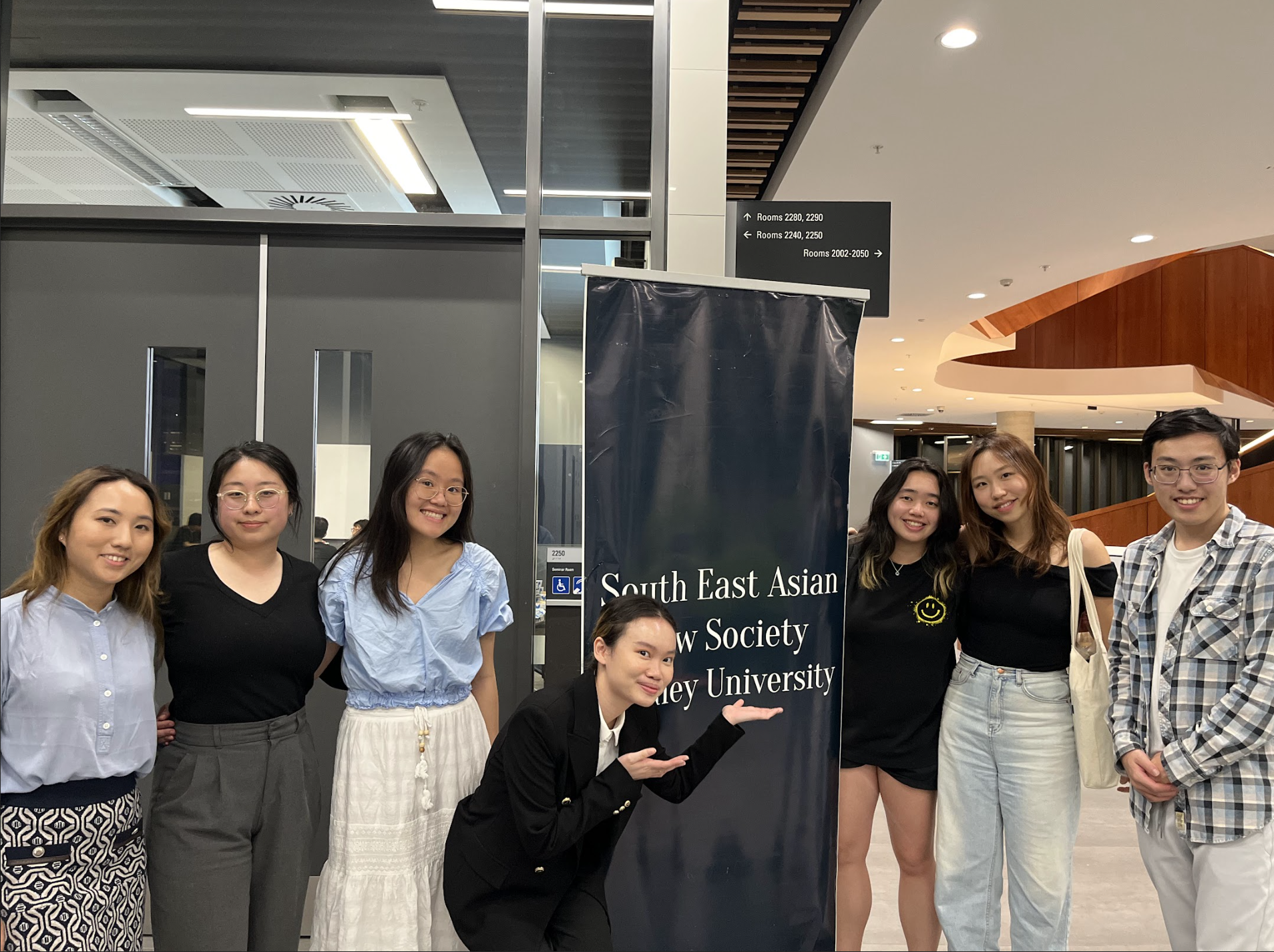 Group of seven young adults posing together at an indoor event, standing beside a navy banner that reads 'South East Asian Law Society Sydney University'.