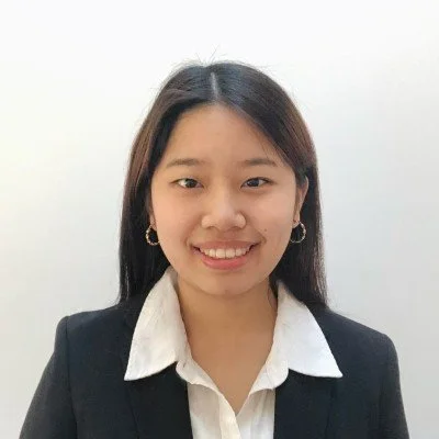 A young woman in a white collared shirt and black blazer smiling at the camera, with long dark hair and hoop earrings, against a plain white background.