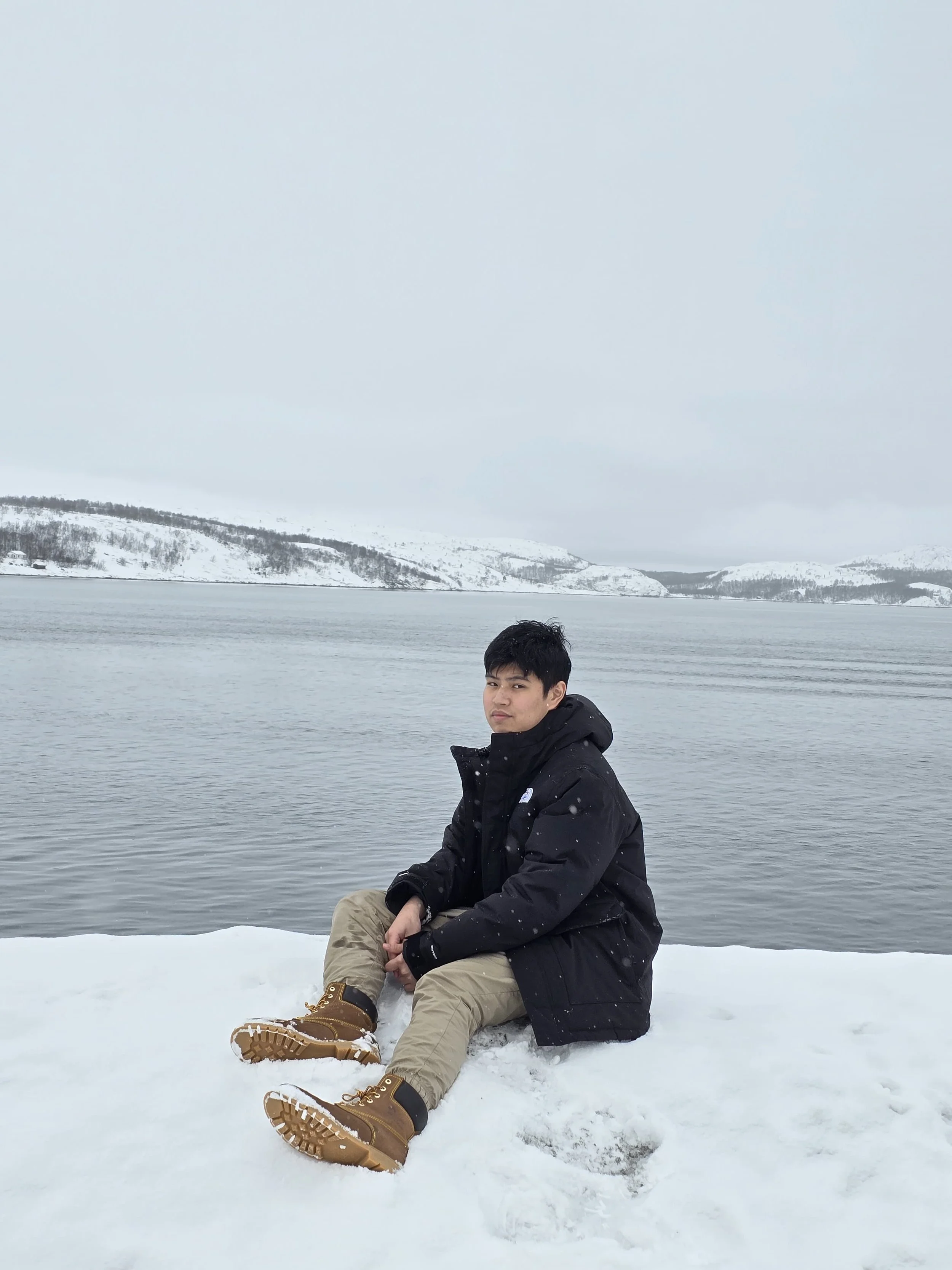 A person sitting on snow near a body of water with snowy hills in the background.