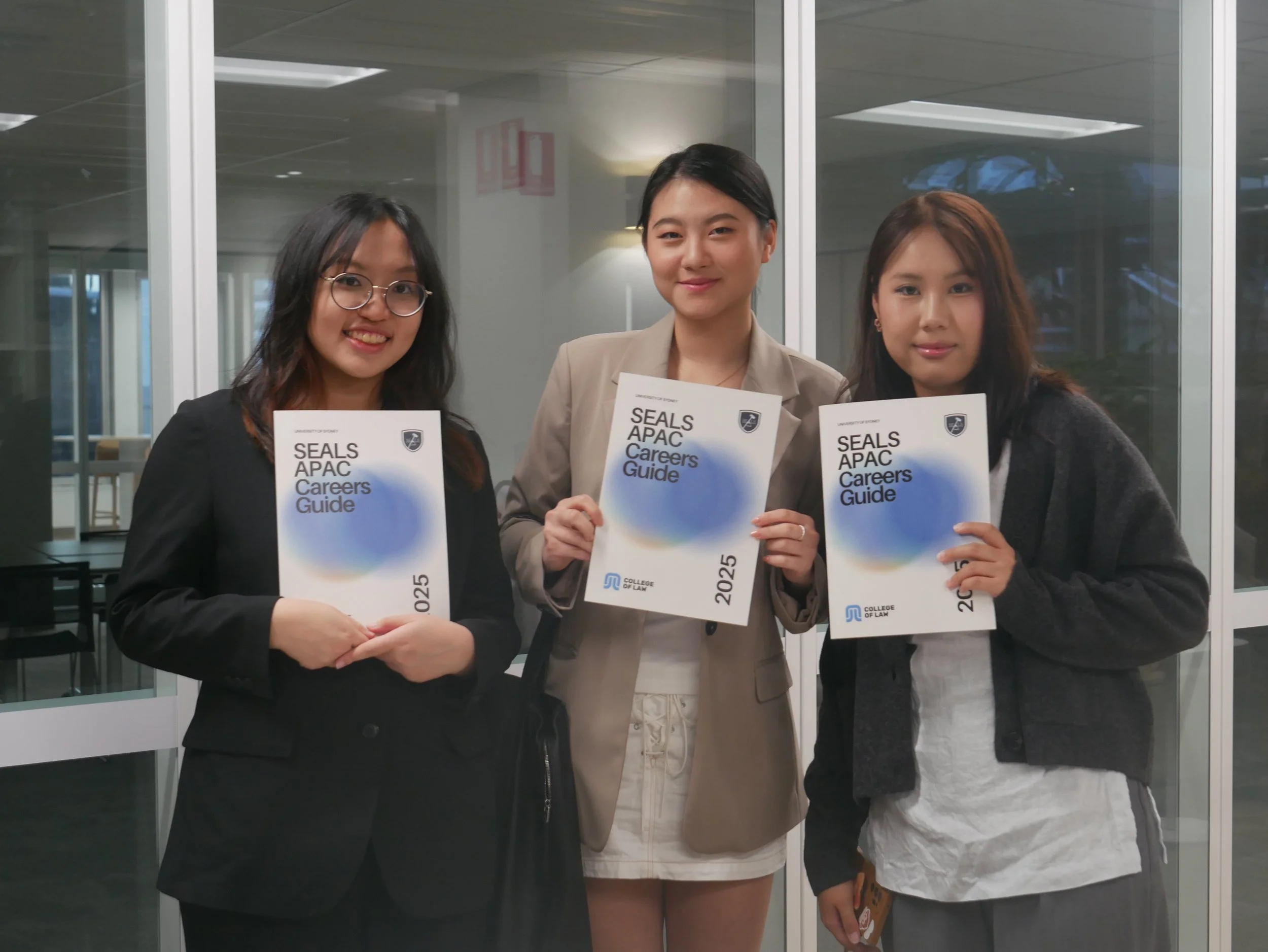 Three young women standing together inside, each holding a booklet titled 'SEALS APAC Careers Guide 2025' from the College of Law, smiling at the camera.