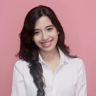 Young woman with long dark hair styled in loose waves, wearing a white shirt, smiling, against a pink background.