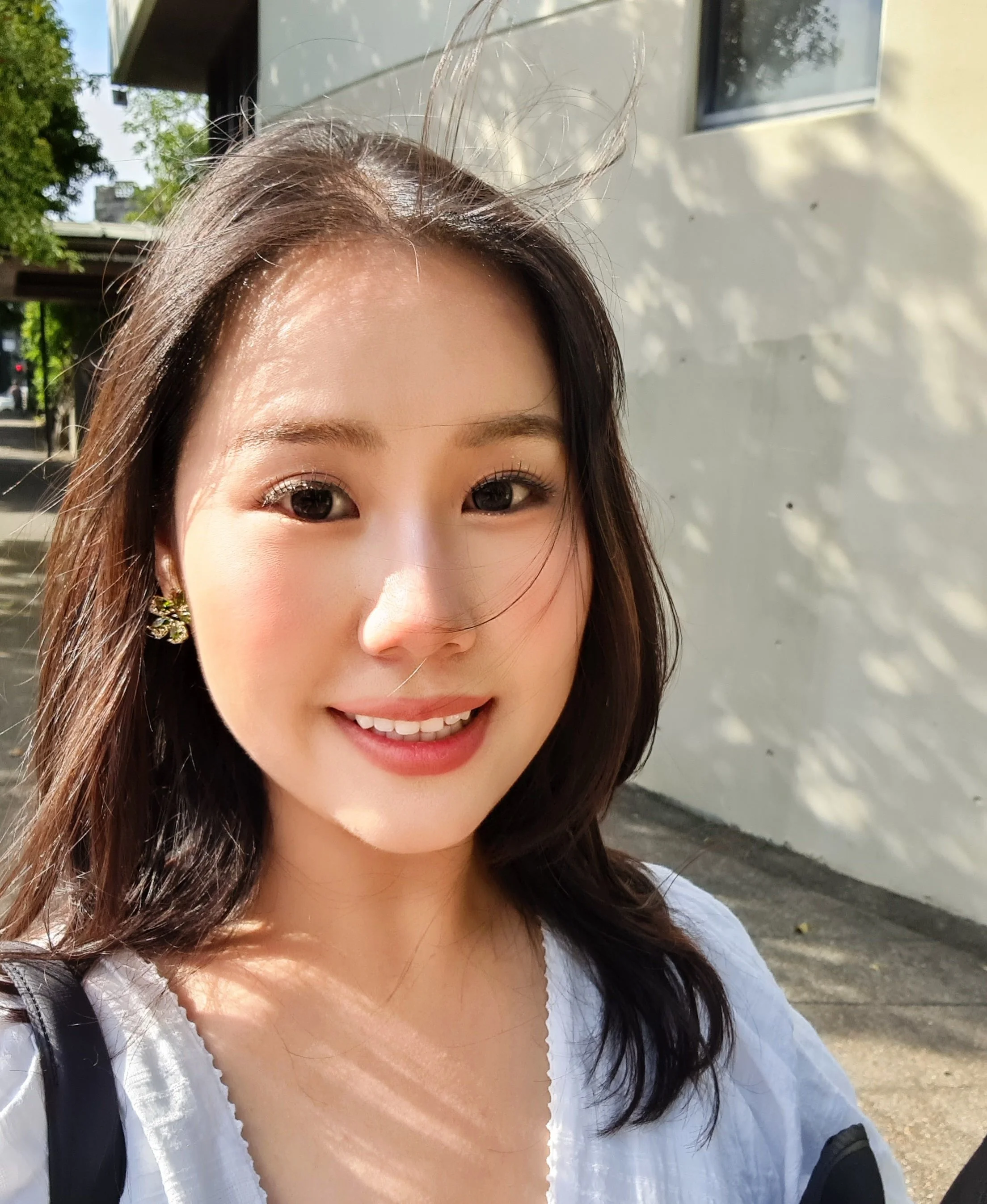 Close-up of a smiling woman with dark brown hair and earrings, outdoors in sunlight with shadows on a wall behind her.