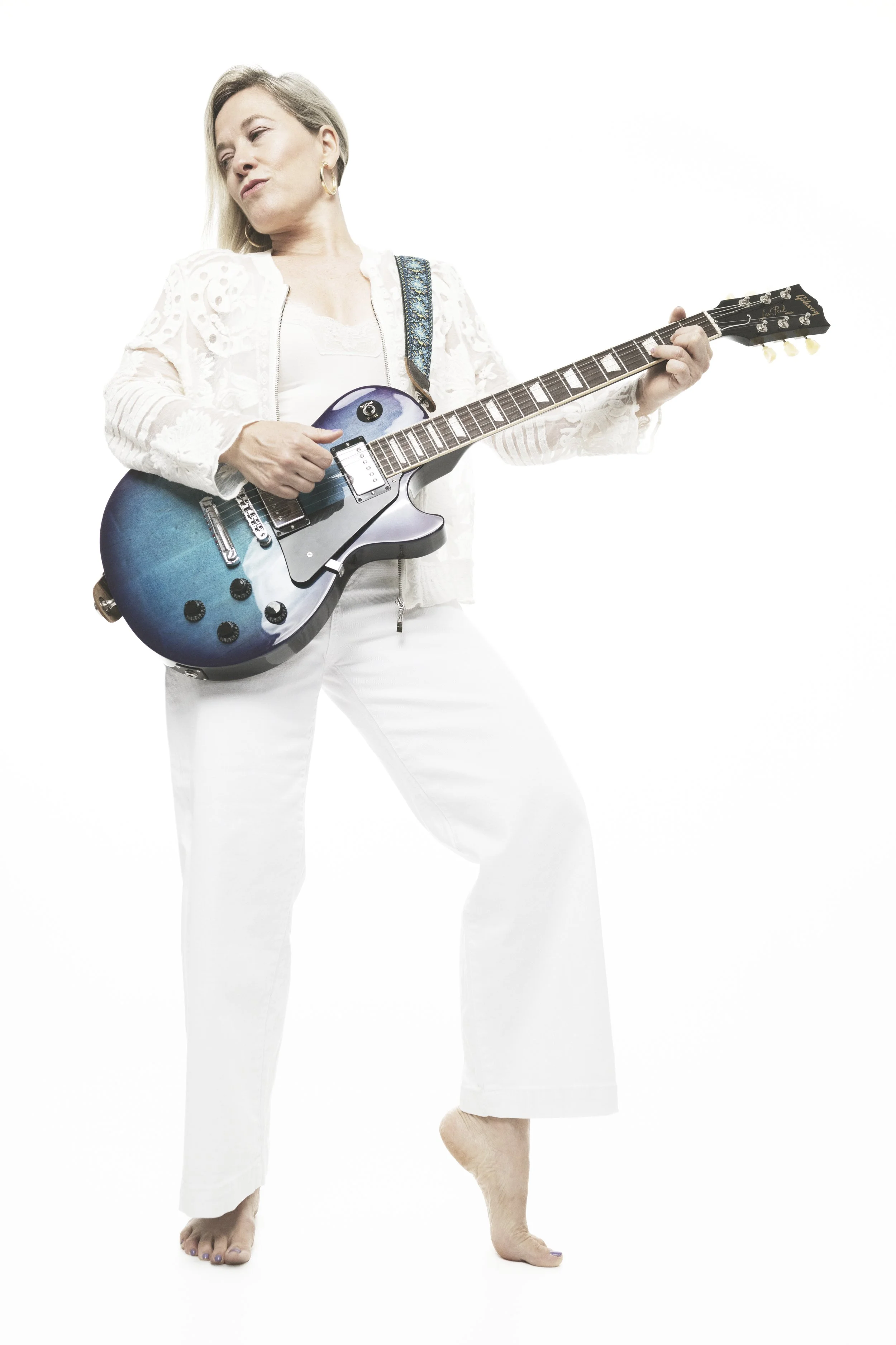 Woman in white pants and top playing electric guitar against a white background, with her feet visible.
