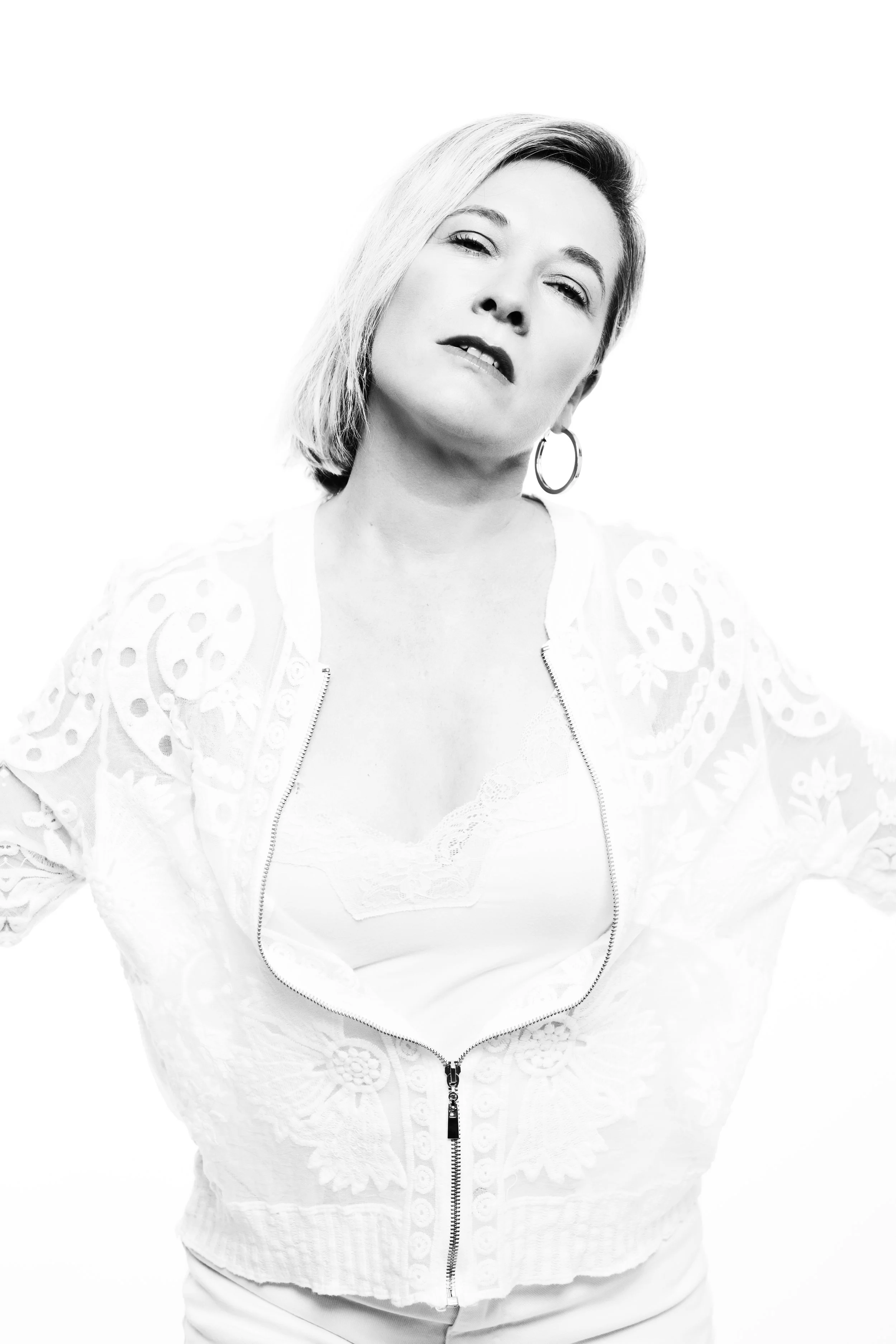 Black and white photo of a woman with styled short hair, wearing a zipper-front embroidered jacket and hoop earrings, posing with a slightly tilted head and a relaxed facial expression.