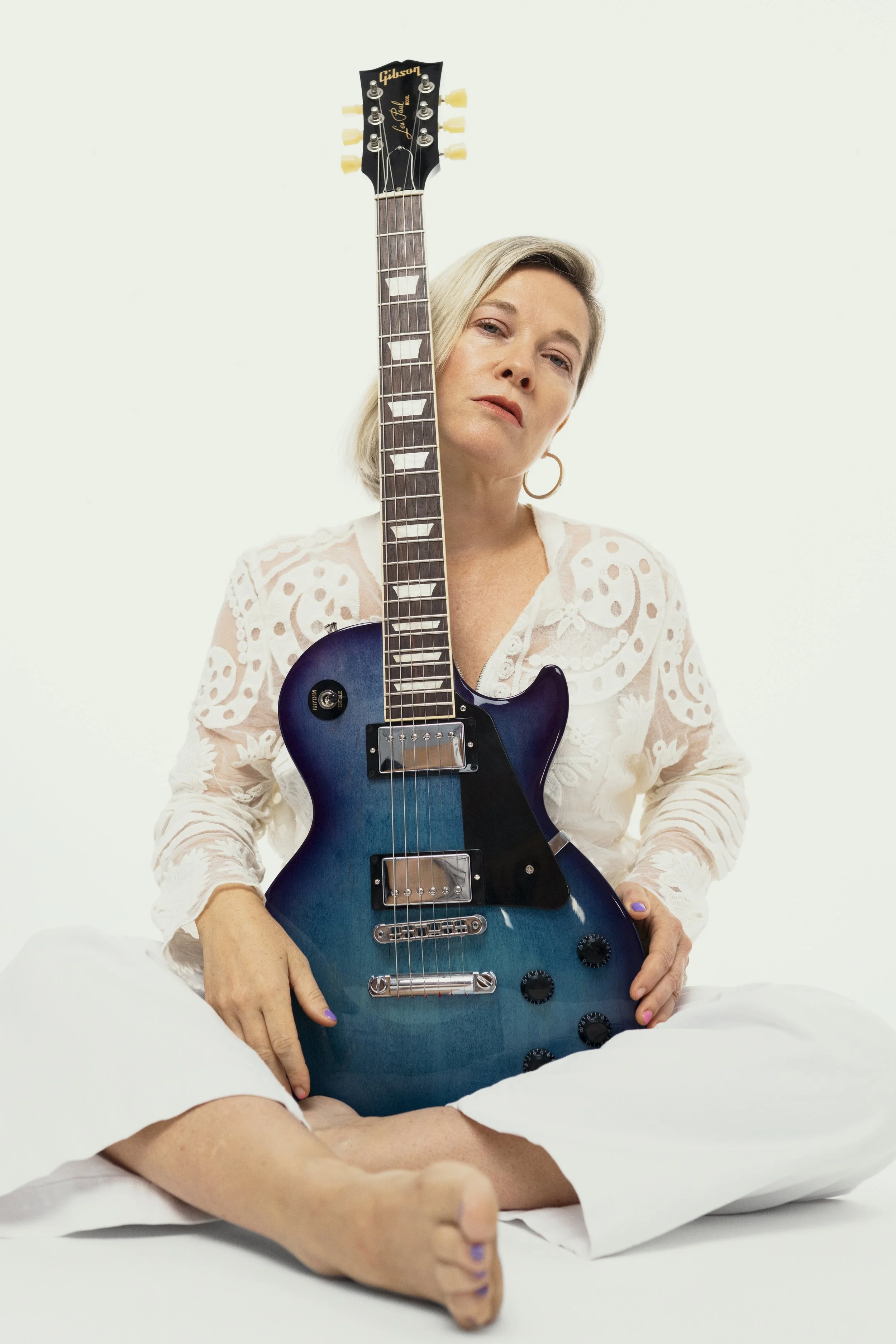 A woman with blond hair holding a blue electric guitar while sitting with her legs crossed, wearing a white embroidered blouse, against a plain white background.