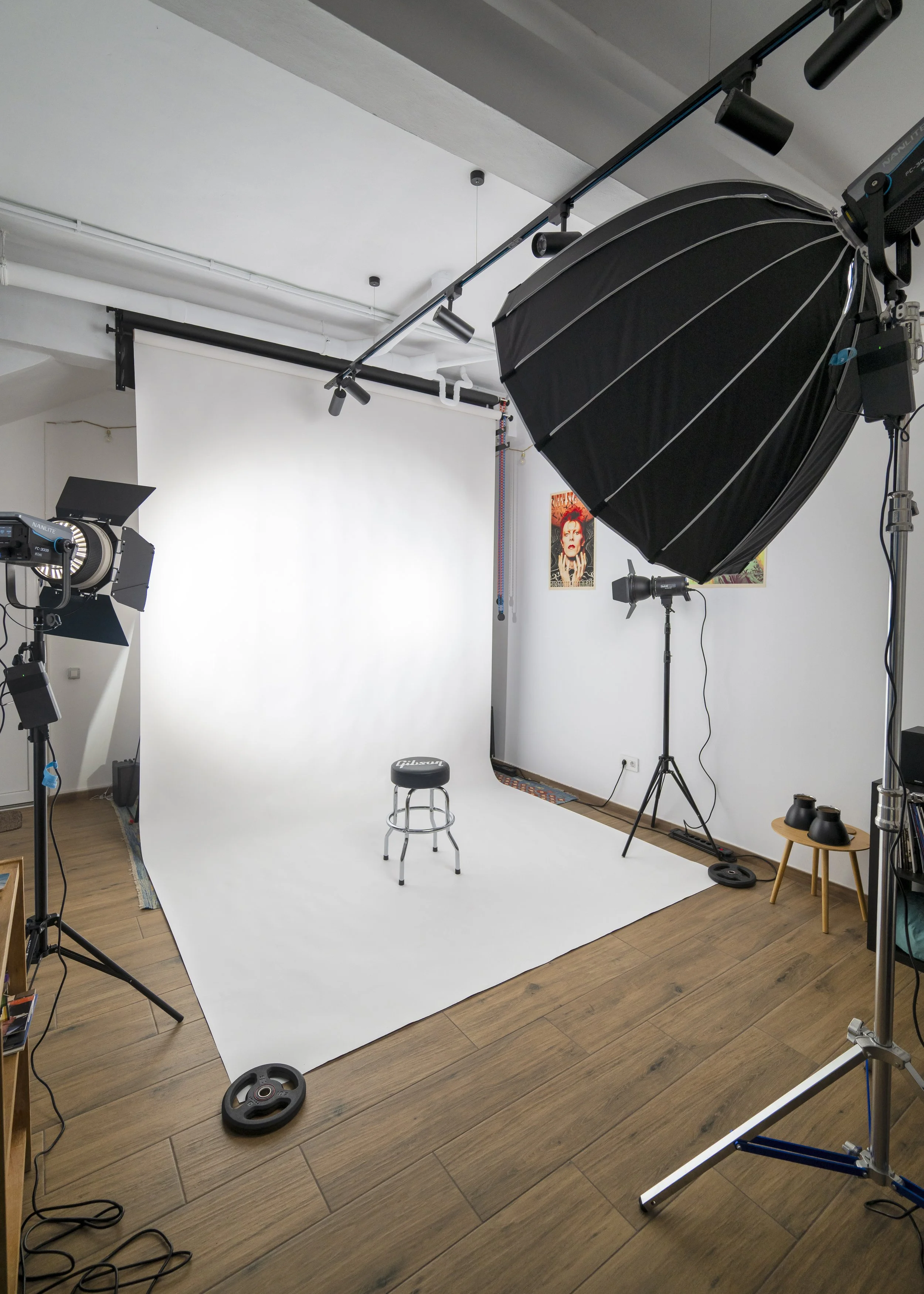 Photography studio with a white backdrop, lighting equipment, a stool, and posters on the wall.