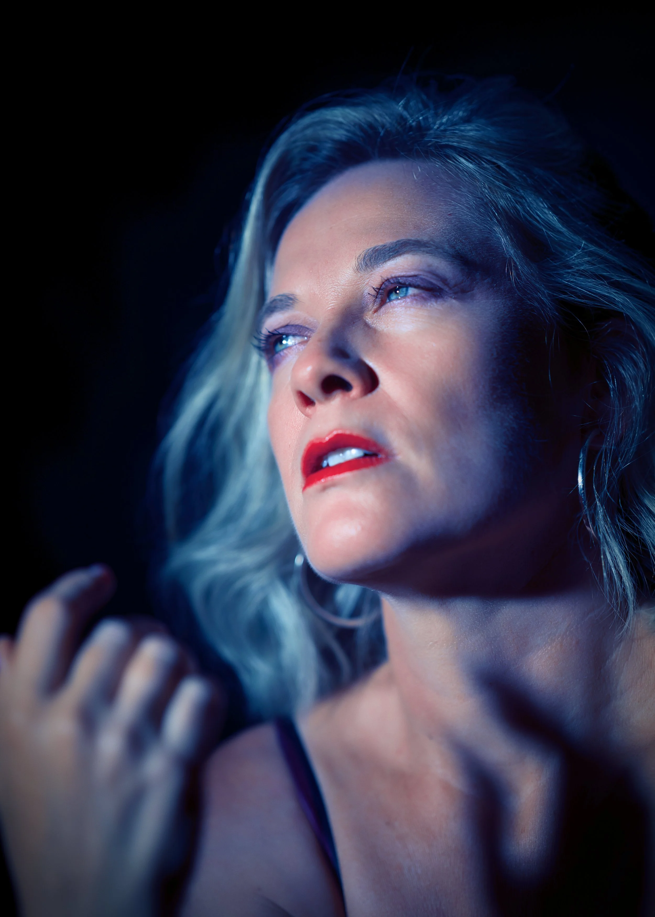 Close-up of a woman with blue and blonde hair, vibrant makeup, and blue eyes, looking upward, with a dark background.