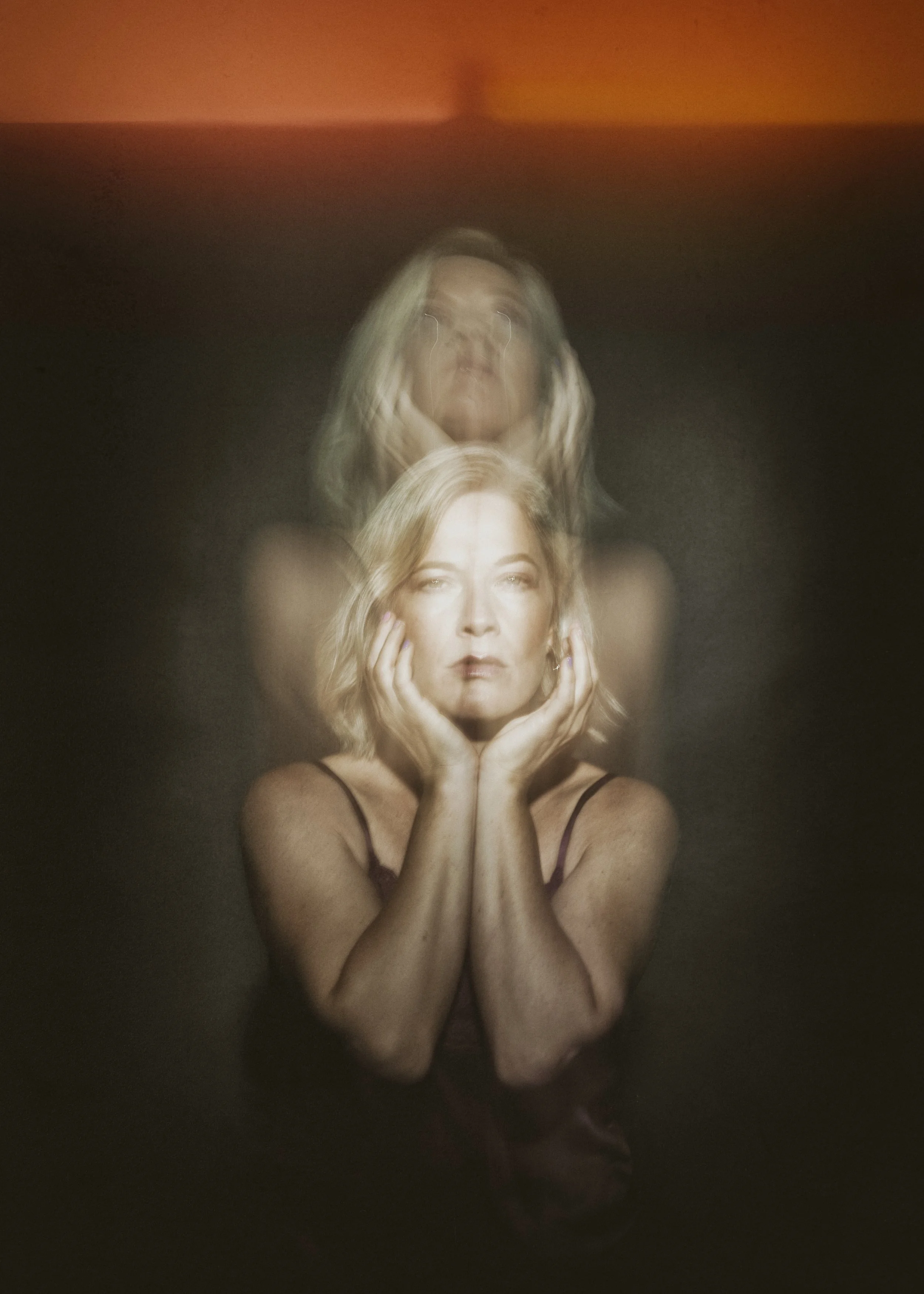 A woman with blonde hair, wearing a black spaghetti strap top, touching her face and looking straight at the camera with her hands framing her face. The image has a double exposure effect with her reflection appearing above her.