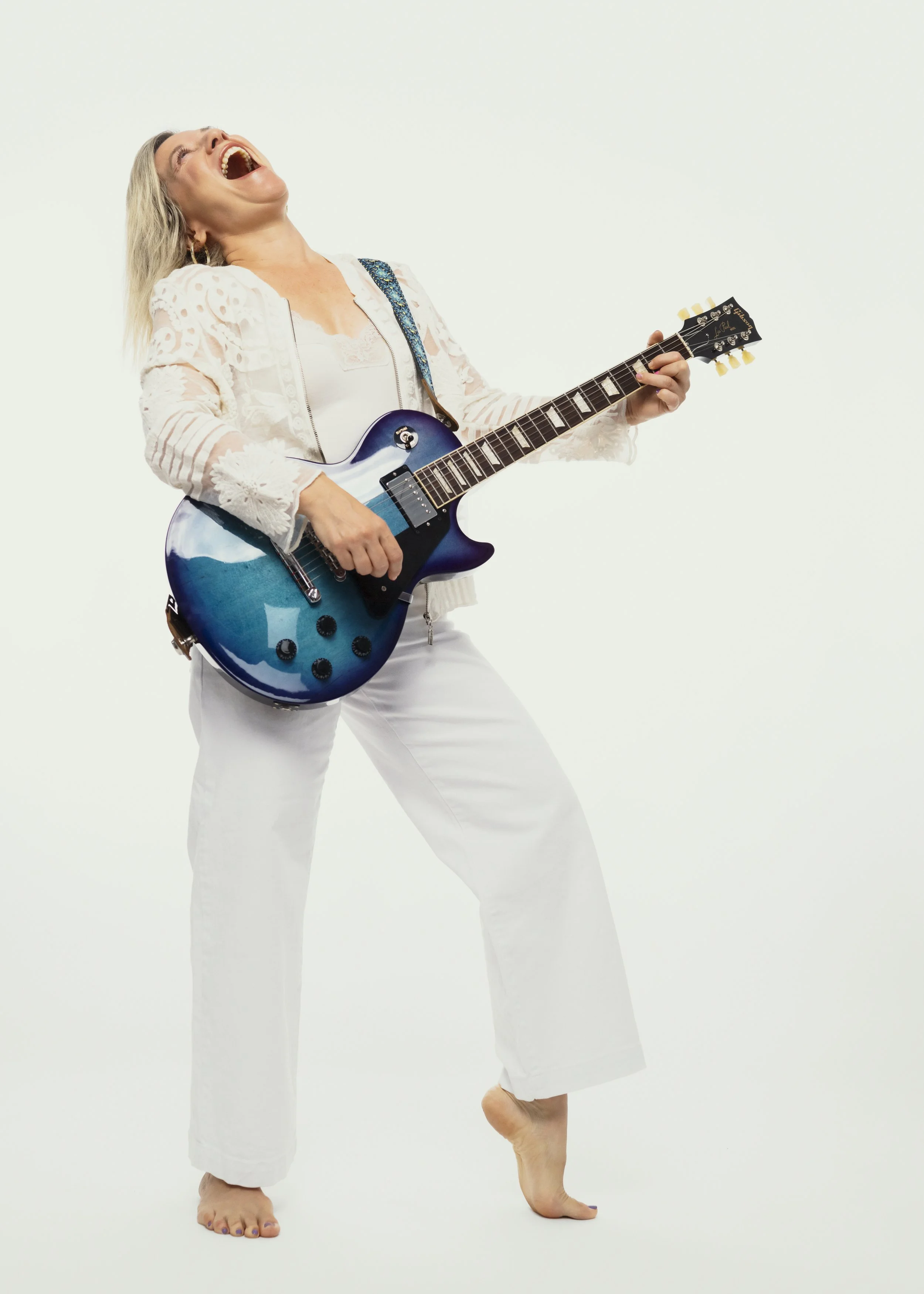 A woman laughing and playing an electric guitar, standing on one foot with the other foot lifted, against a plain white background.