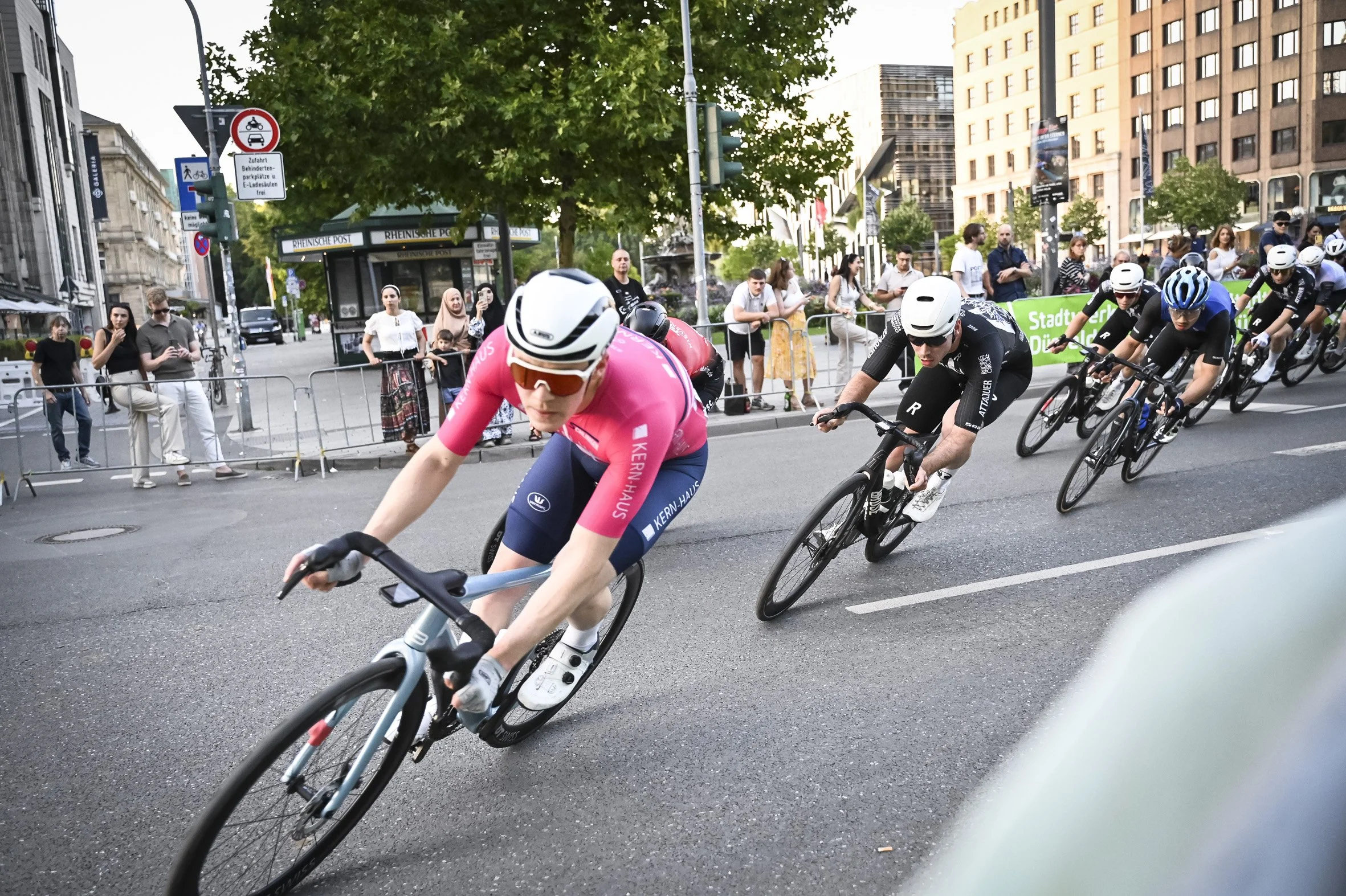 Radsport-Wettbewerb in einer Stadtstraße mit mehreren Fahrern, die um die Kurve fahren, während Zuschauer im Hintergrund zuschauen.