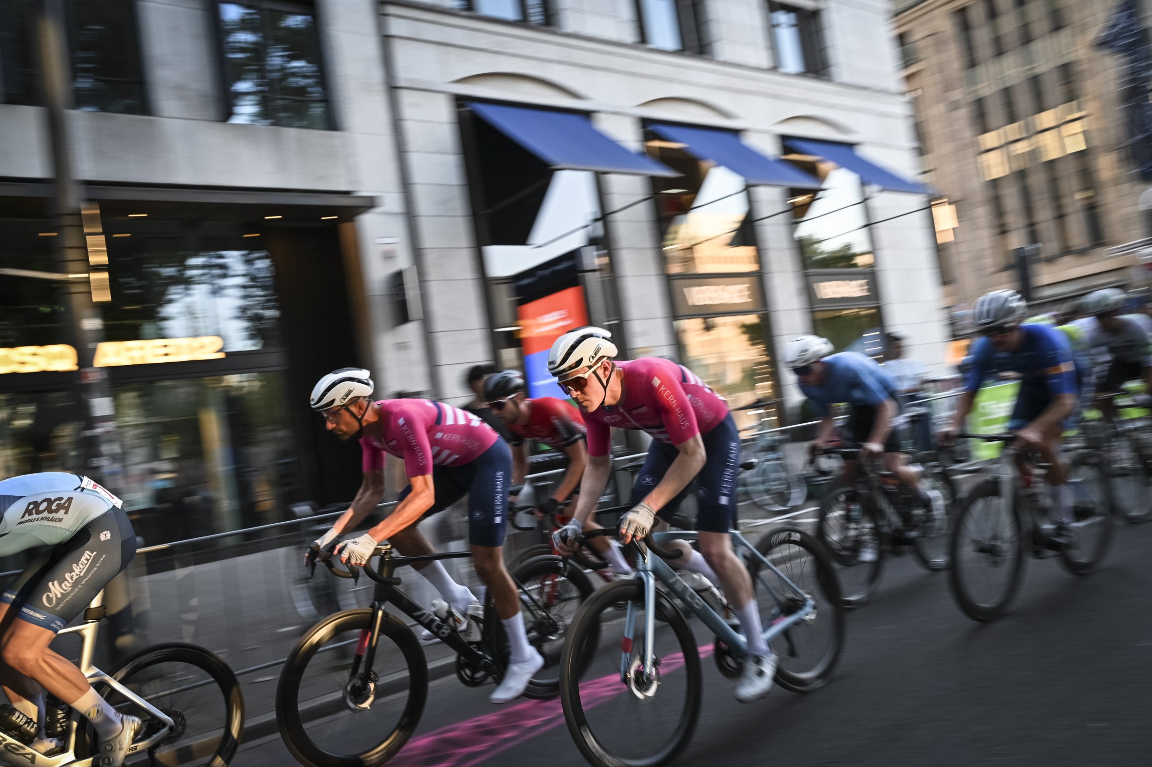 Radfahrer in einem Stadtrennen, alle tragen helme und sportive Kleidung, auf einer urbanen Straße mit Gebäuden im Hintergrund.