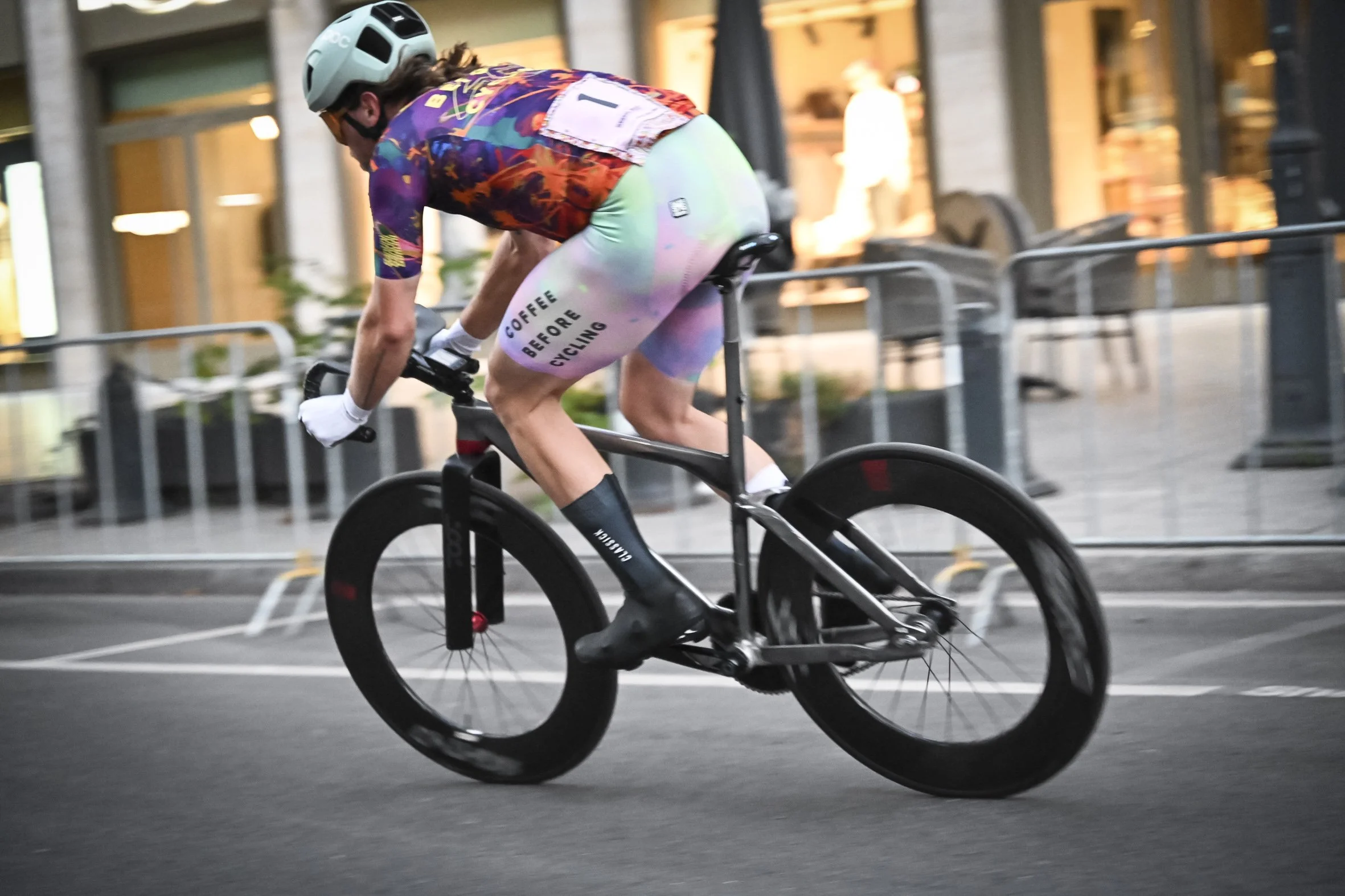 Fahrradfahrer trägt einen Helm und bunte Radsportkleidung, fährt auf einer Straße in einer Stadt bei Dämmerung.