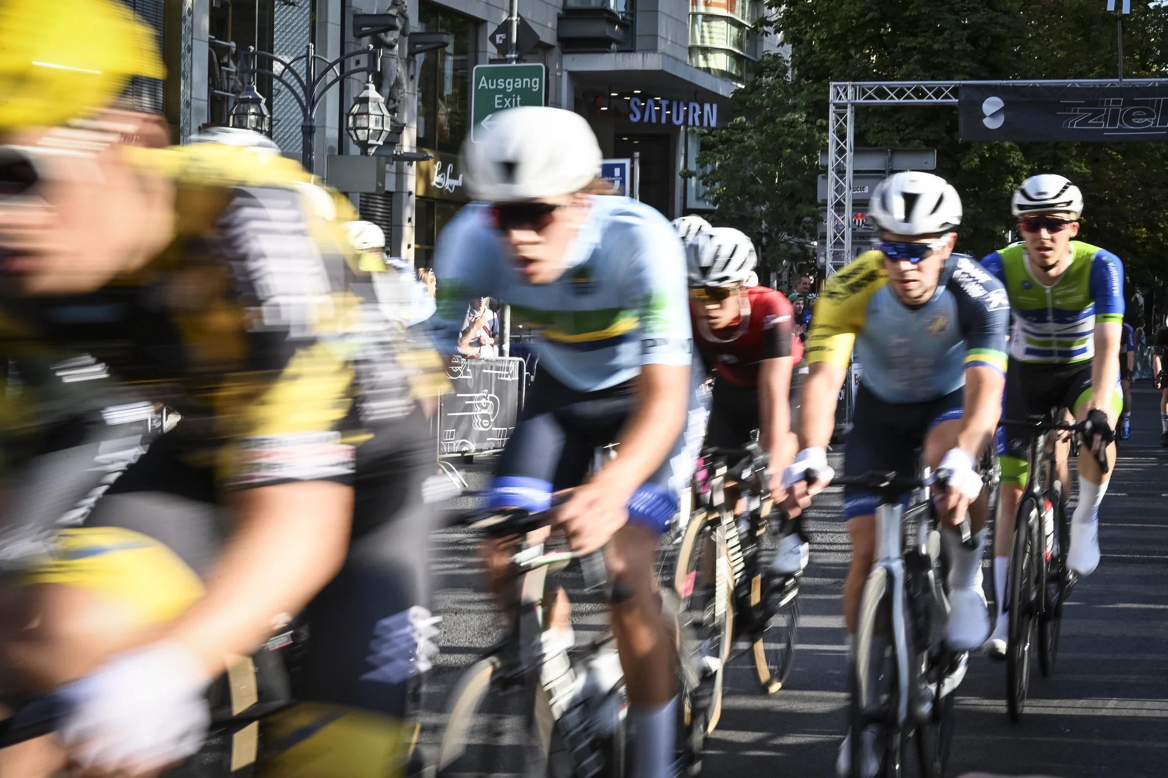 Radfahrer bei einem Rennen auf einer Stadtstraße, einige tragen Helme und Sonnenbrillen, im Hintergrund sind Geschäfte und ein Schild mit der Aufschrift 'Saturn' sichtbar.