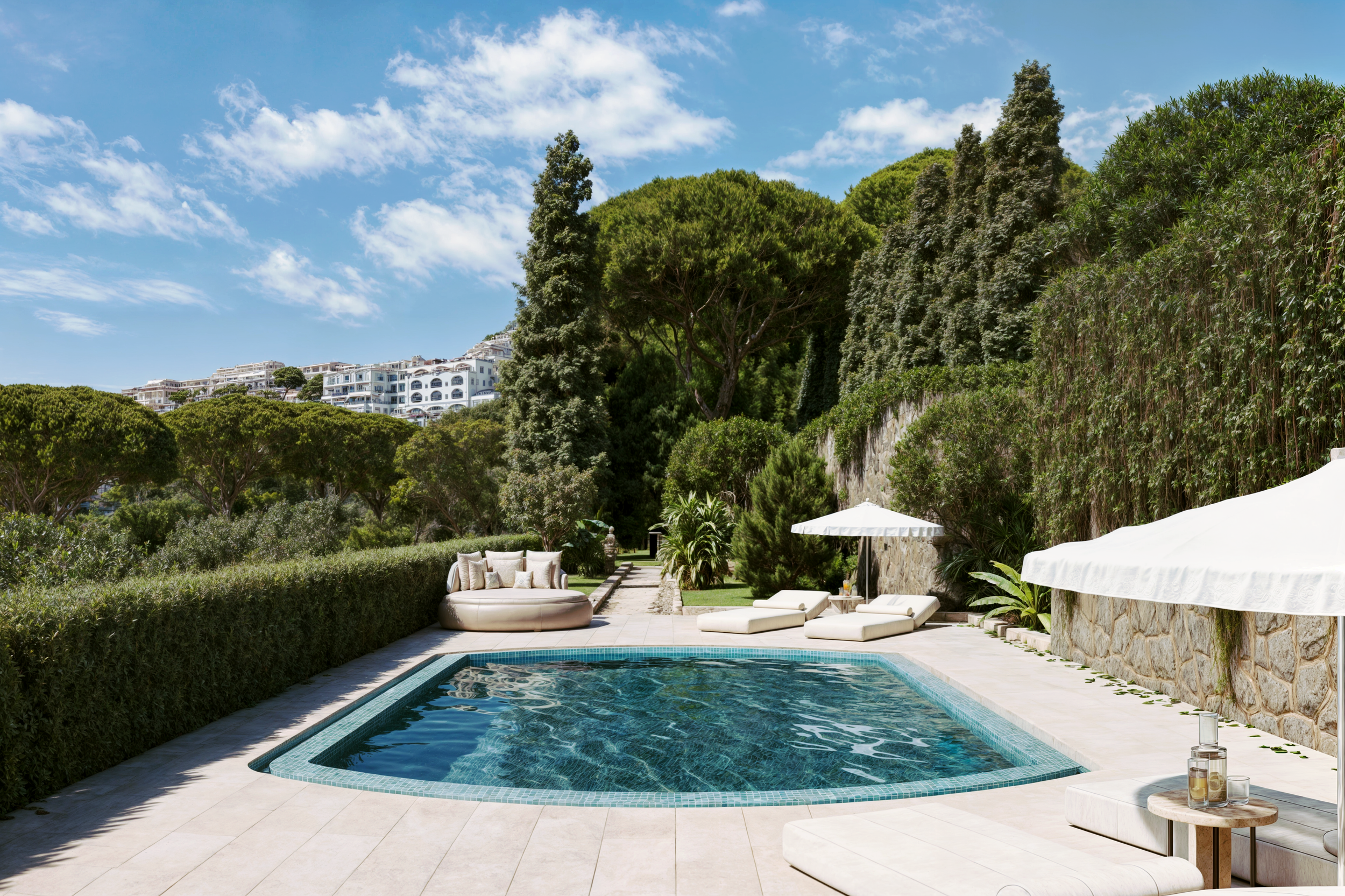 Piscina ao ar livre com espreguiçadeiras, guarda-sol e área de descanso, cercada por vegetação e árvores, com moradias no topo de uma colina ao fundo, sob céu azul com nuvens.
