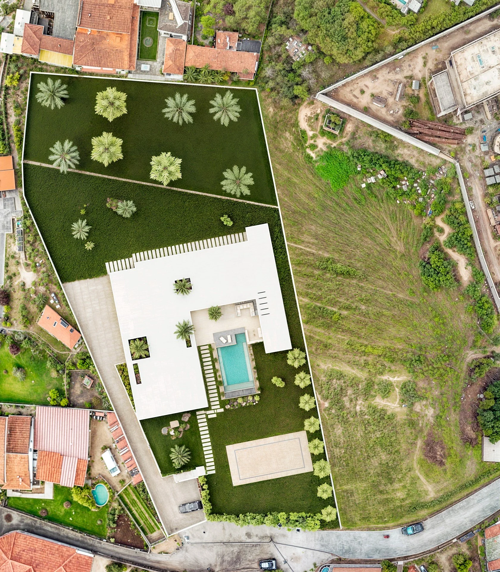 Imagem aérea de uma residência com piscina, quadra de esportes, área de lazer, jardim e árvores ao redor, em uma região com casas vizinhas e terreno vazio.