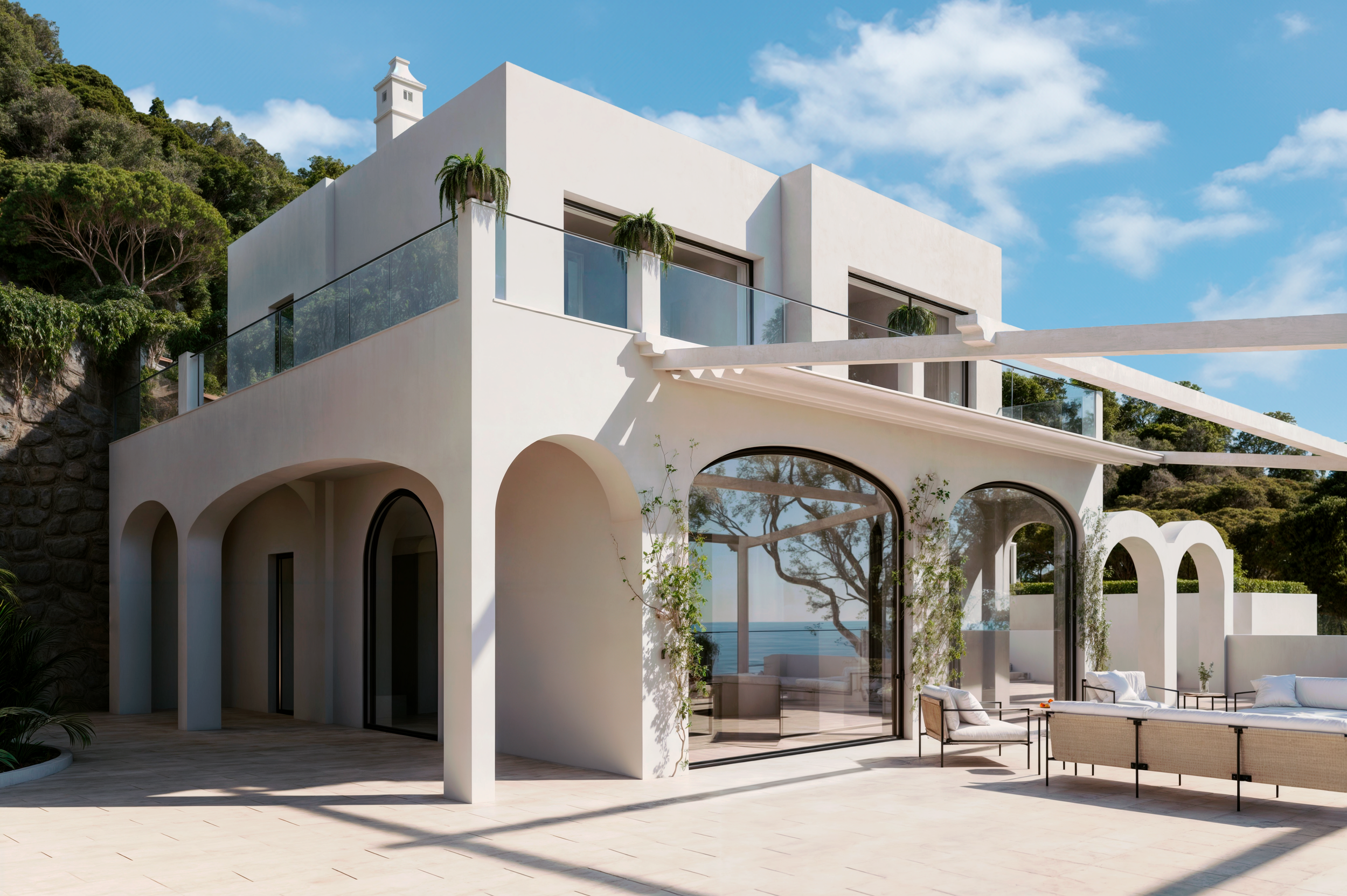 Residência moderna branca com grandes janelas de vidro, varanda com guarda-corpo de vidro, decoração externa com mobília de descanso e plantas, situada em área com vegetação e vista para o mar, sob céu claro com nuvens.