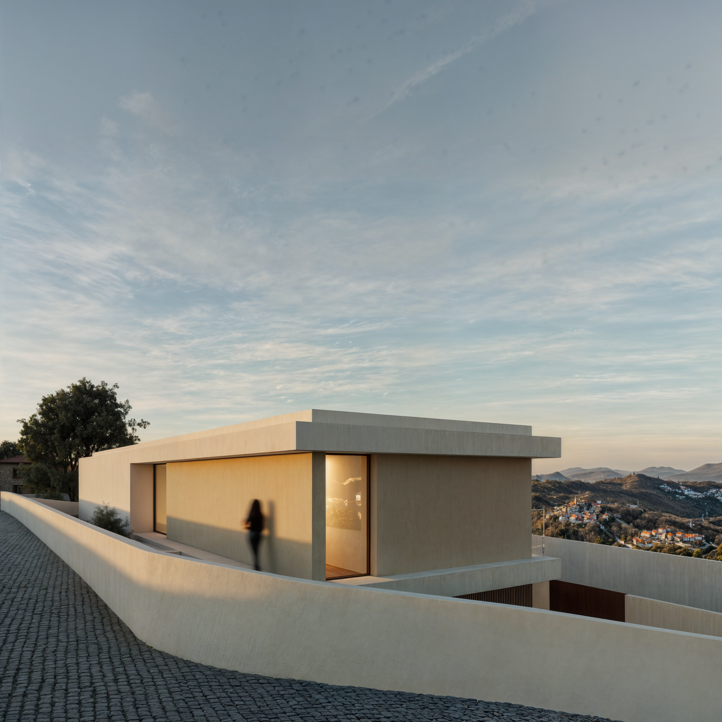 Edifício moderno de cor clara com janelas de vidro, localizado em uma área elevada com vista para uma paisagem de montanhas e vilarejos, ao entardecer.
