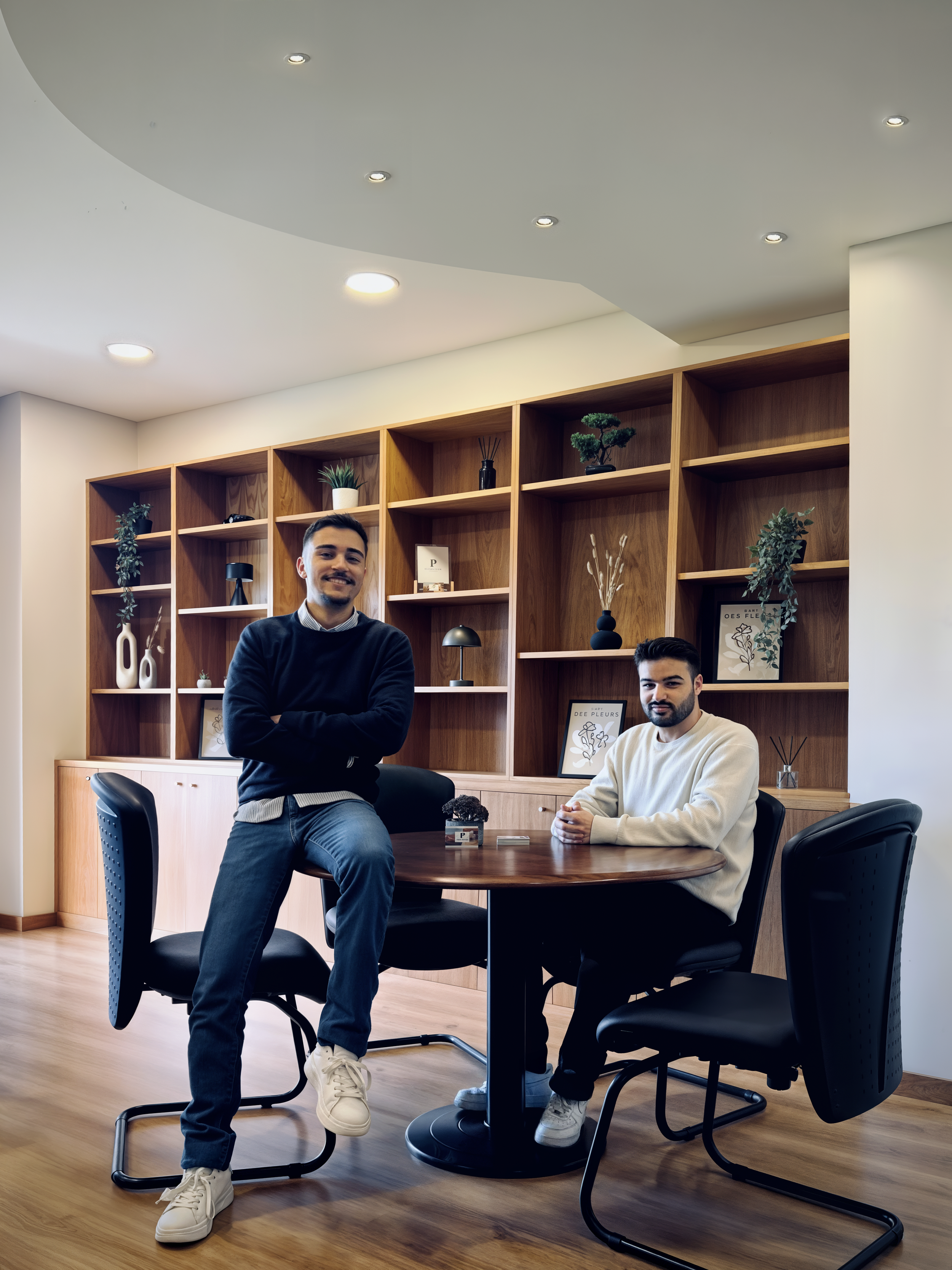 Dois jovens homens em uma sala de reuniões com estantes de madeira ao fundo, decoradas com plantas, quadros e objetos decorativos, um deles de pé ao lado da mesa e o outro sentado.