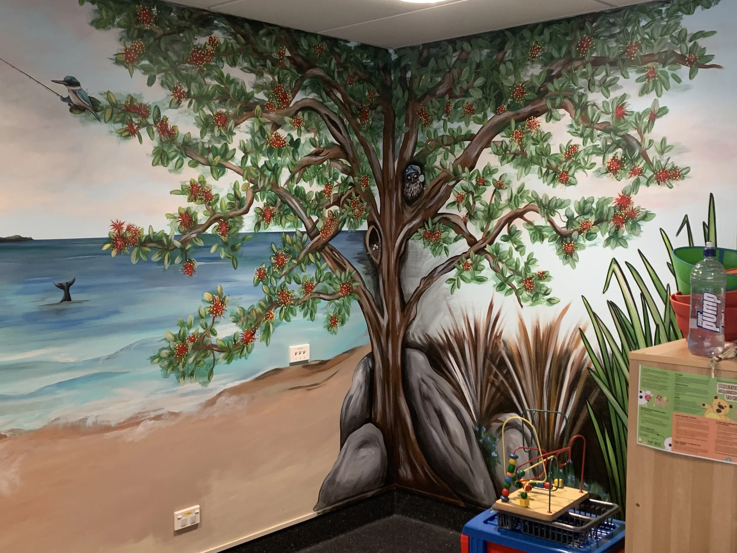 Colorful mural of a beach scene with a large flowering tree, ocean waves, and birds on the wall of a children's playroom, with toys and supplies nearby.