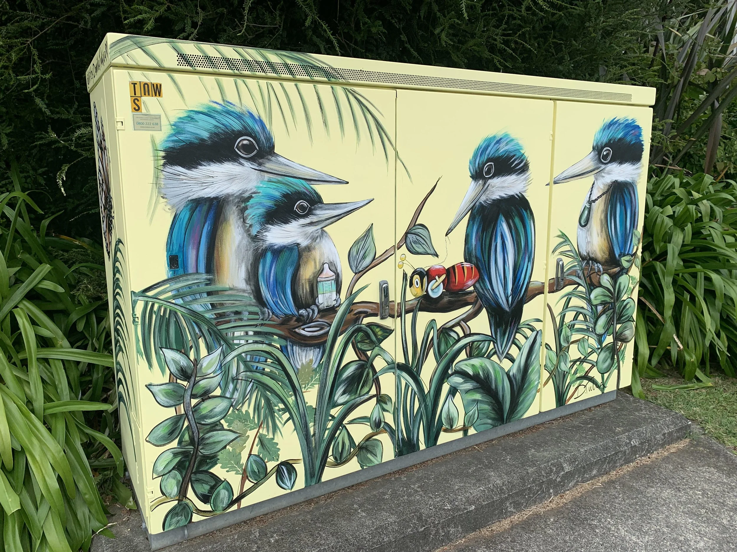 Colorful street art of four kingfisher birds with blue and black feathers, perched among green tropical plants on a utility box.