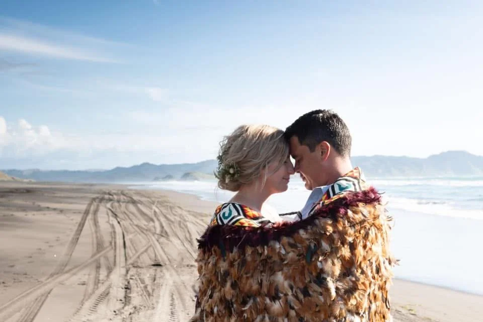 Dannika Tukua and her husband hugging on Kawhia beach, with ocean waves and distant mountains in the background, under a clear blue sky.