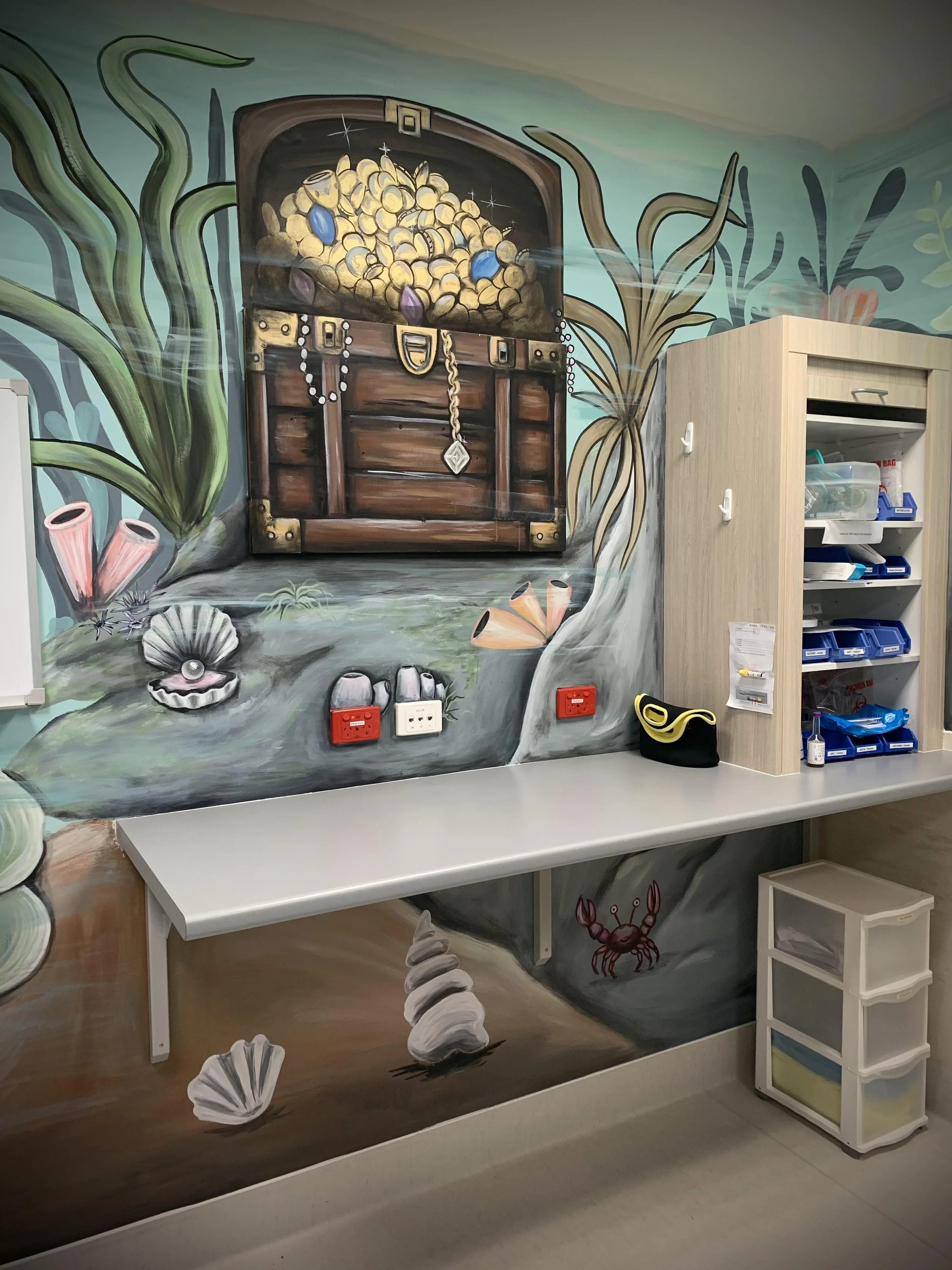 Colorful underwater-themed mural with a treasure chest full of gold coins, seashells, coral, seaweed, and a crab painted on the wall behind a white countertop and storage cabinet.