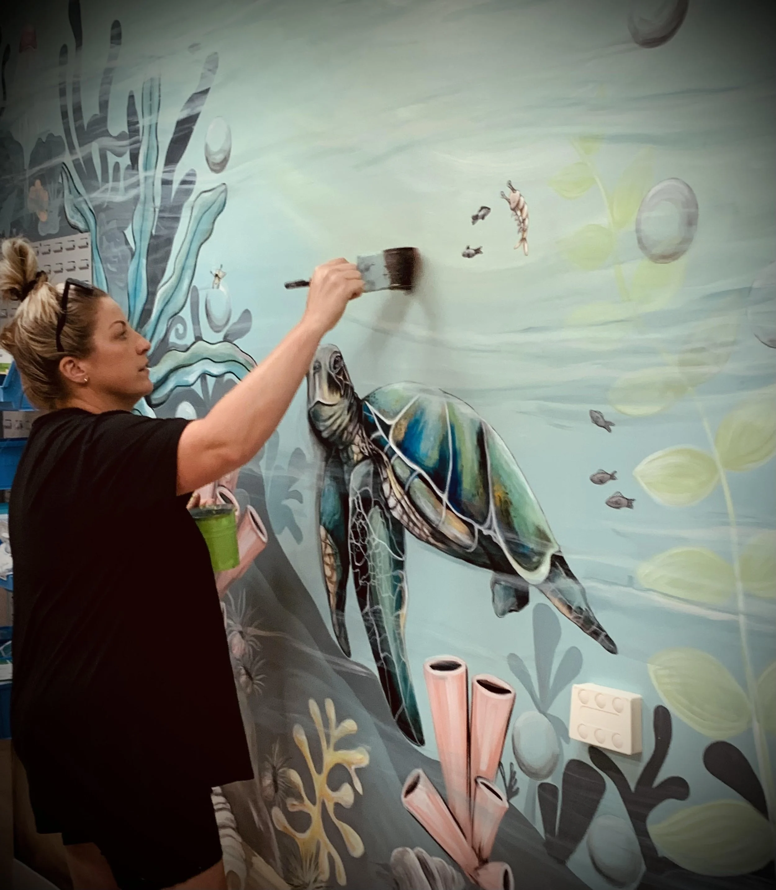 Dannika Tukua painting a mural of an underwater scene featuring a sea turtle, fish, coral, and seaweed on a wall.