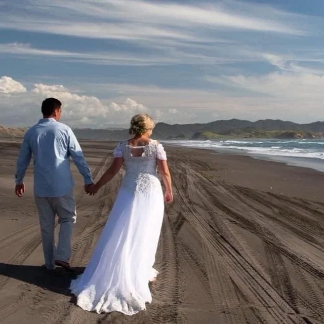 A man and woman in wedding attire holding hands and walking on a sandy beach with ocean waves, mountains in the background, under a partly cloudy sky.