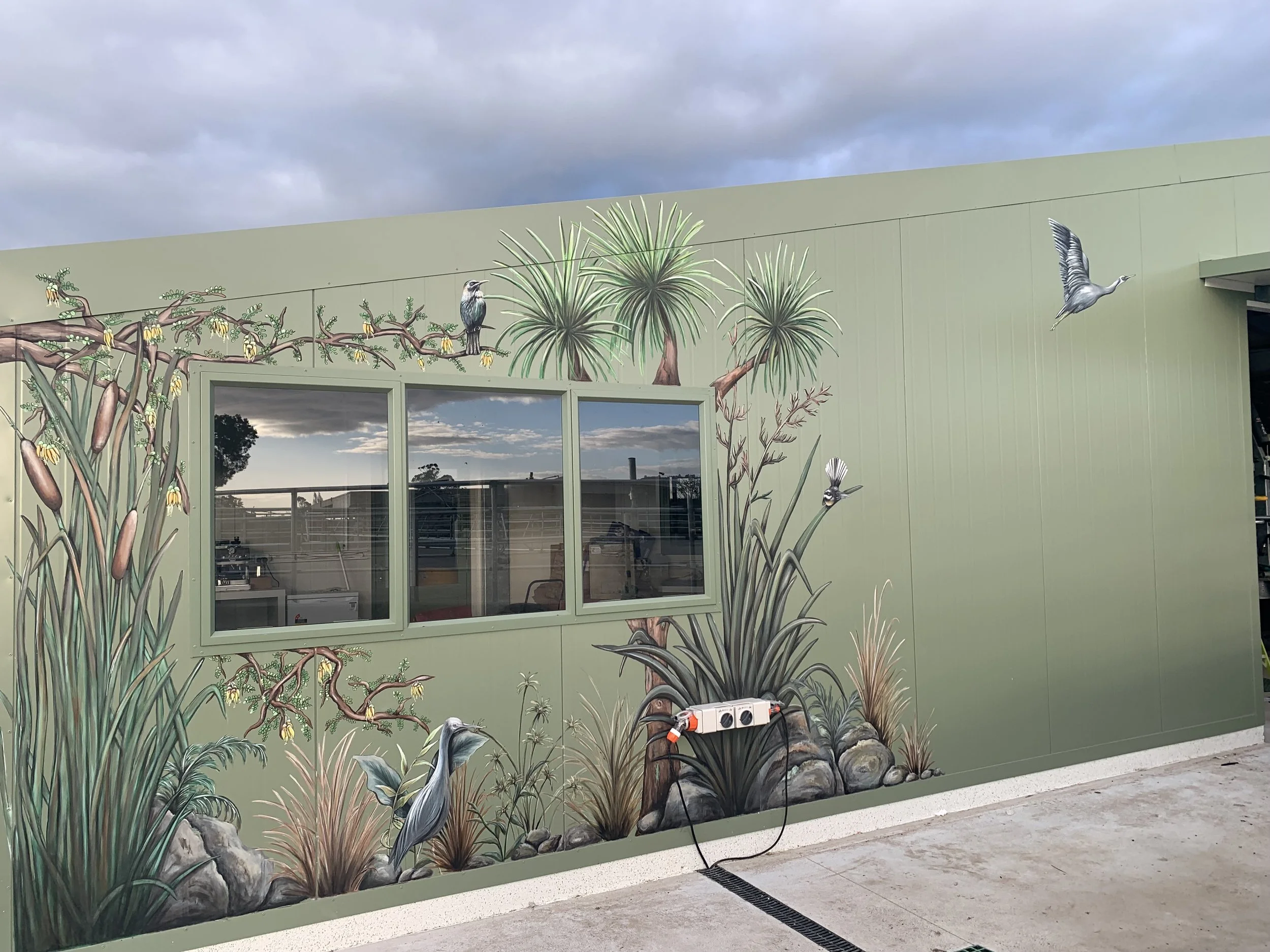 Murals of plants, birds, and animals painted on a green exterior wall near a window, with critters including birds and a fish, against a cloudy sky.
