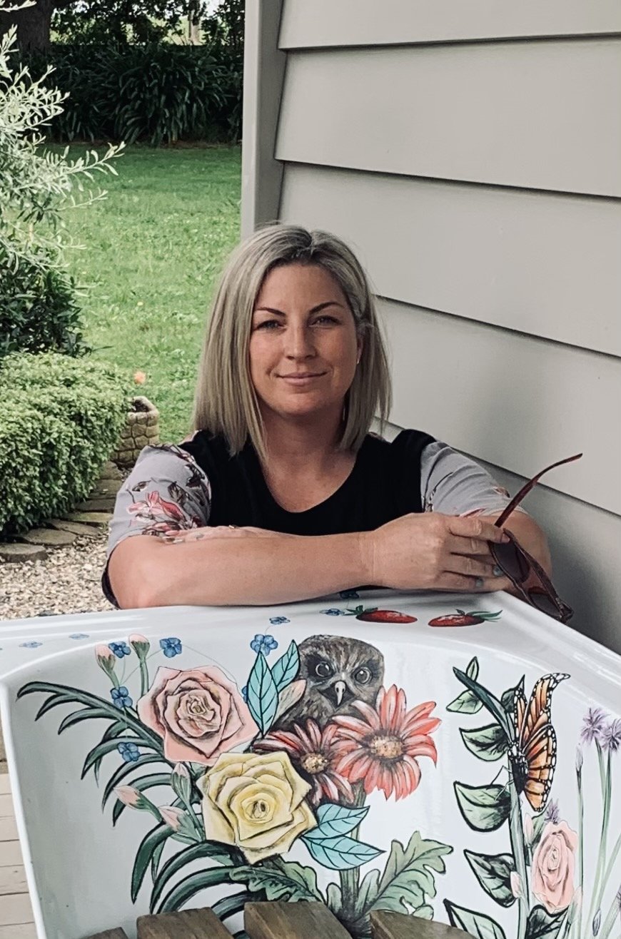 A woman with shoulder-length blonde hair sitting at a table outdoors, leaning on her arm, with a slight smile. The table has a painted design of flowers, a bird, and a butterfly. Background shows green grass, bushes, and a house siding.