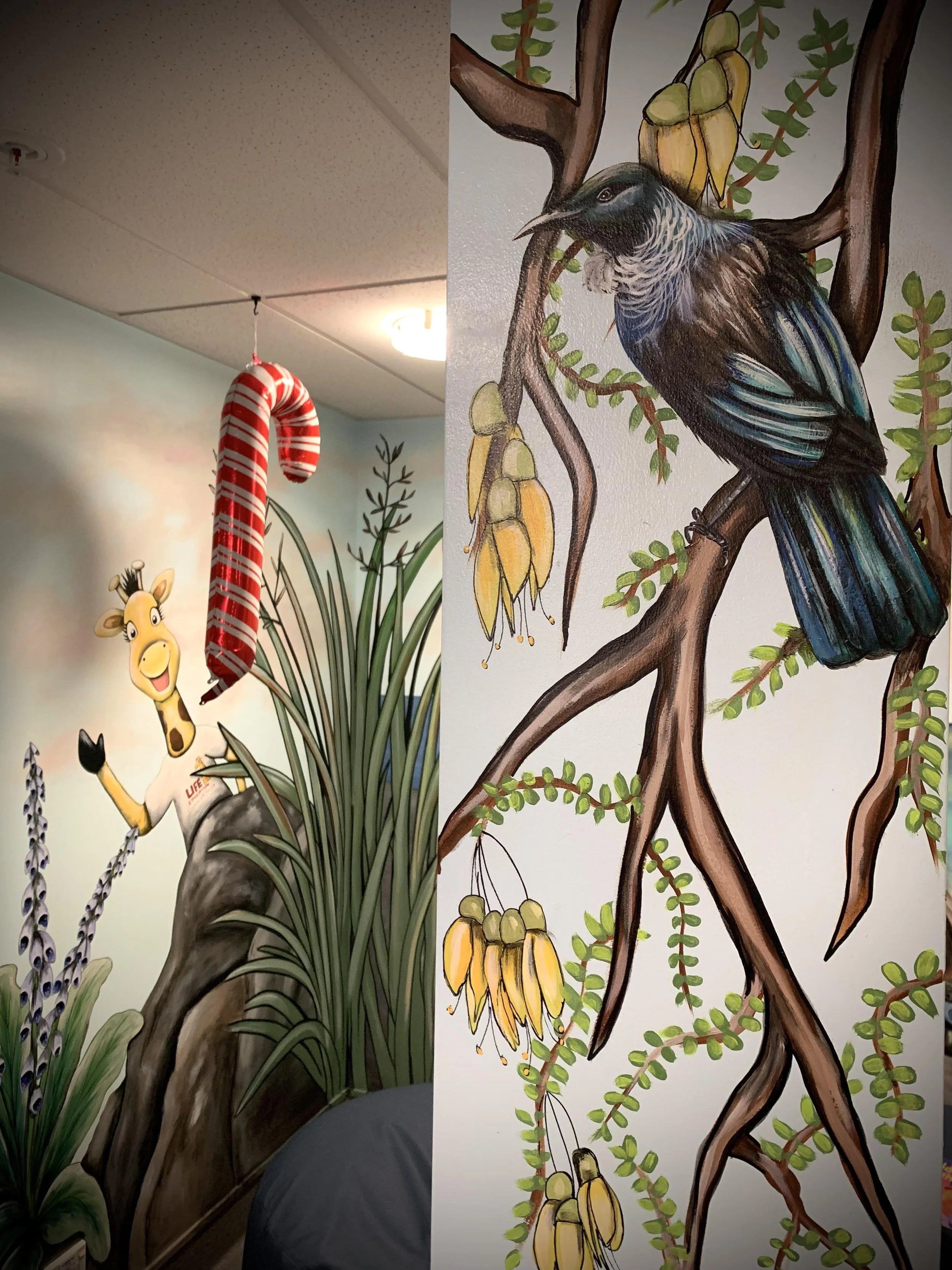 Colorful indoor mural featuring a cartoon giraffe, a bird perched on a tree branch, and various plants and flowers.