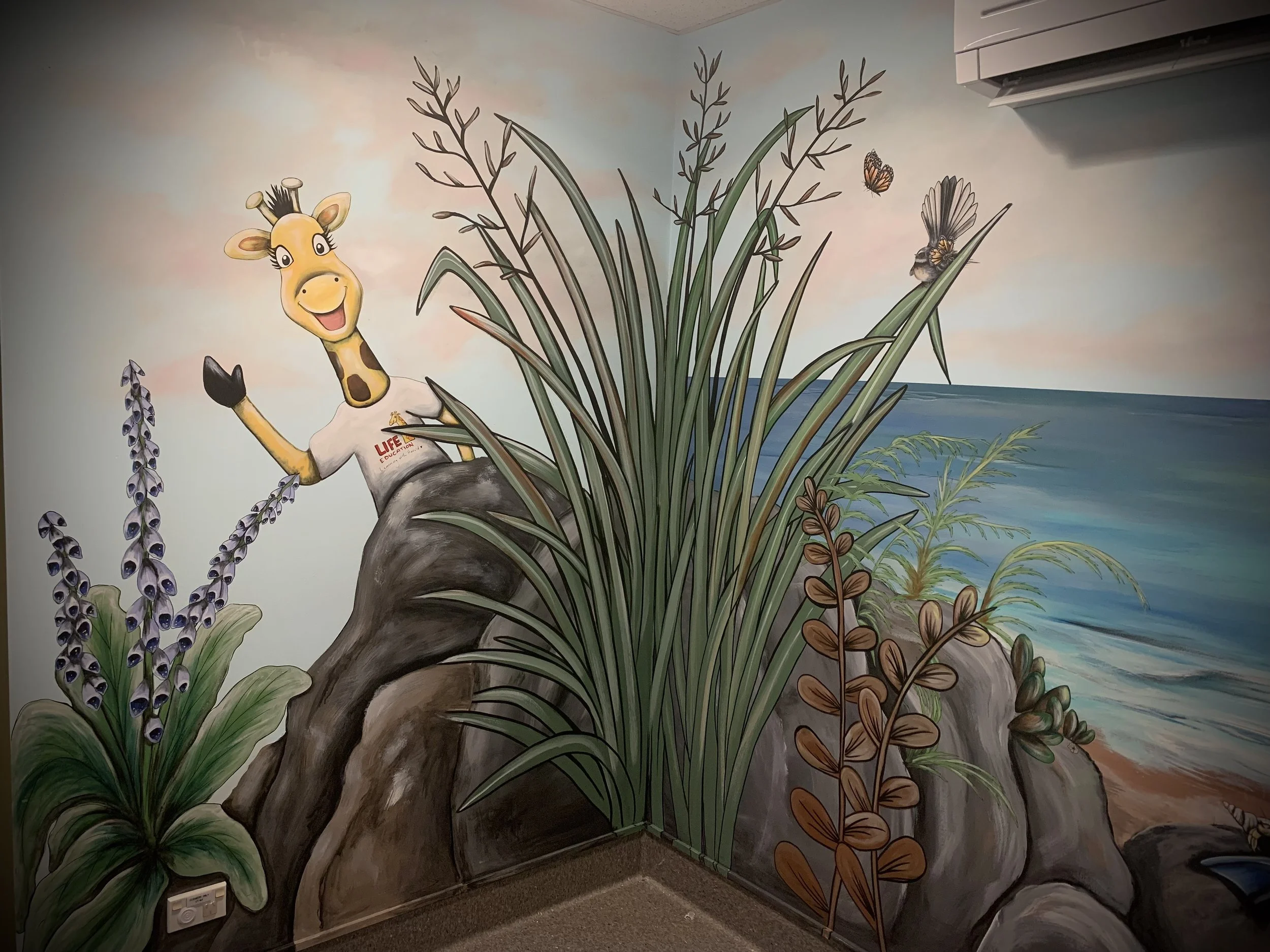 Colorful mural featuring a cheerful giraffe wearing a shirt, standing on a rock among various plants, with a background of the ocean and sky, including butterflies and a bird.