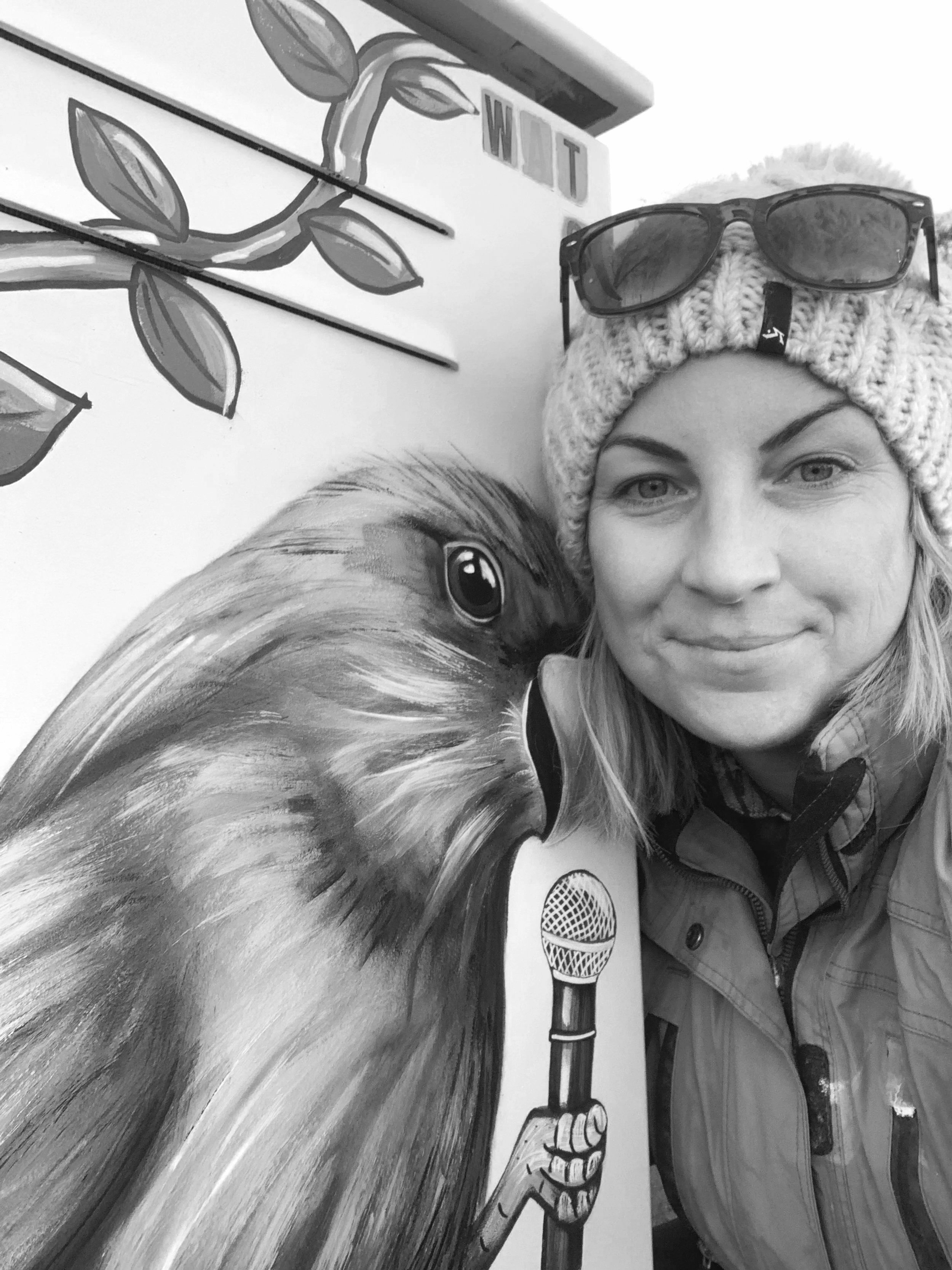 A black and white photo of Dannika Tukua wearing a knit hat and sunglasses on her forehead, taking a selfie with a mural of a Kakariki bird holding a microphone.