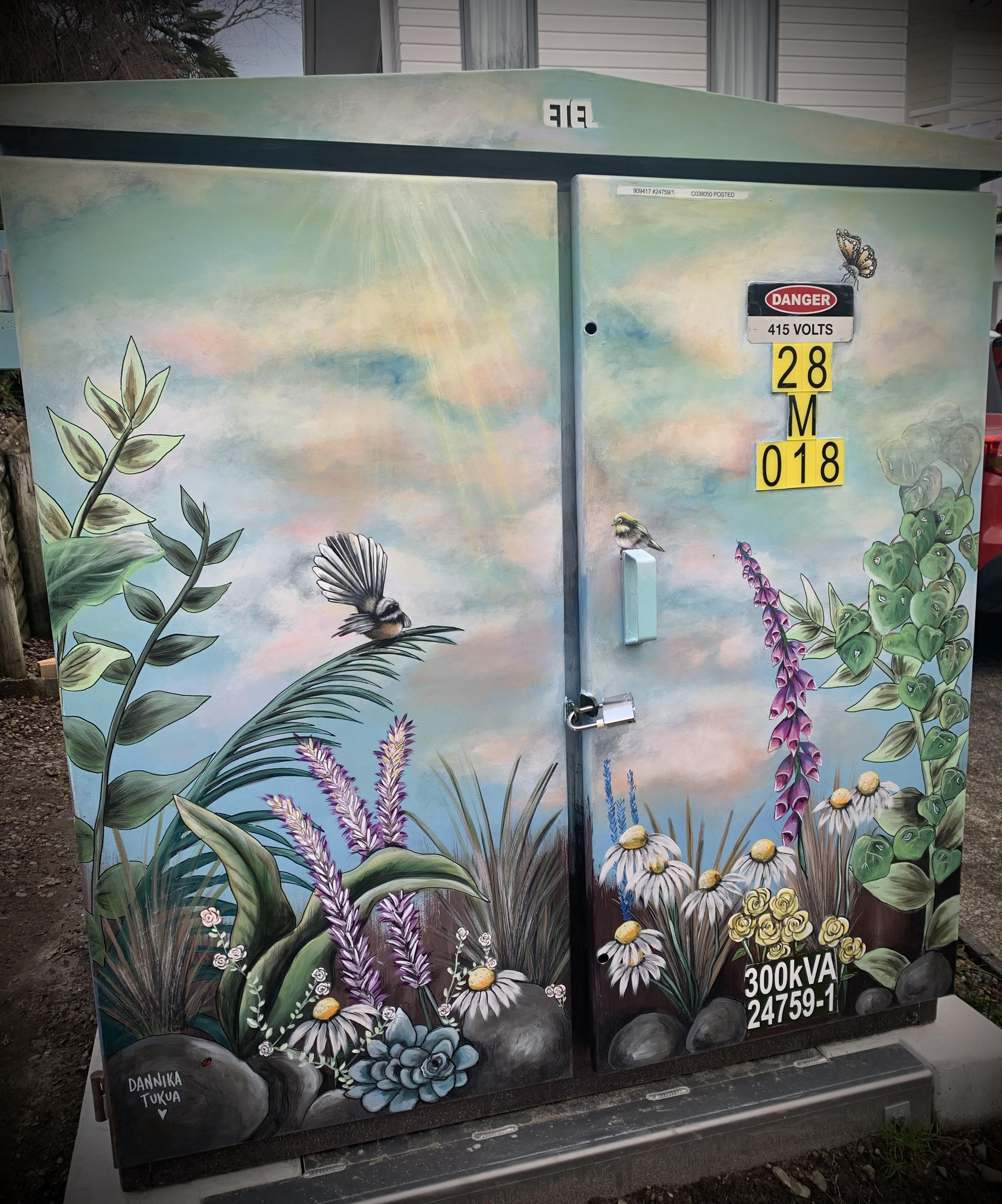 Decorated electrical box with nature-themed mural featuring grass, flowers, and two birds, along with warning labels and electrical information.