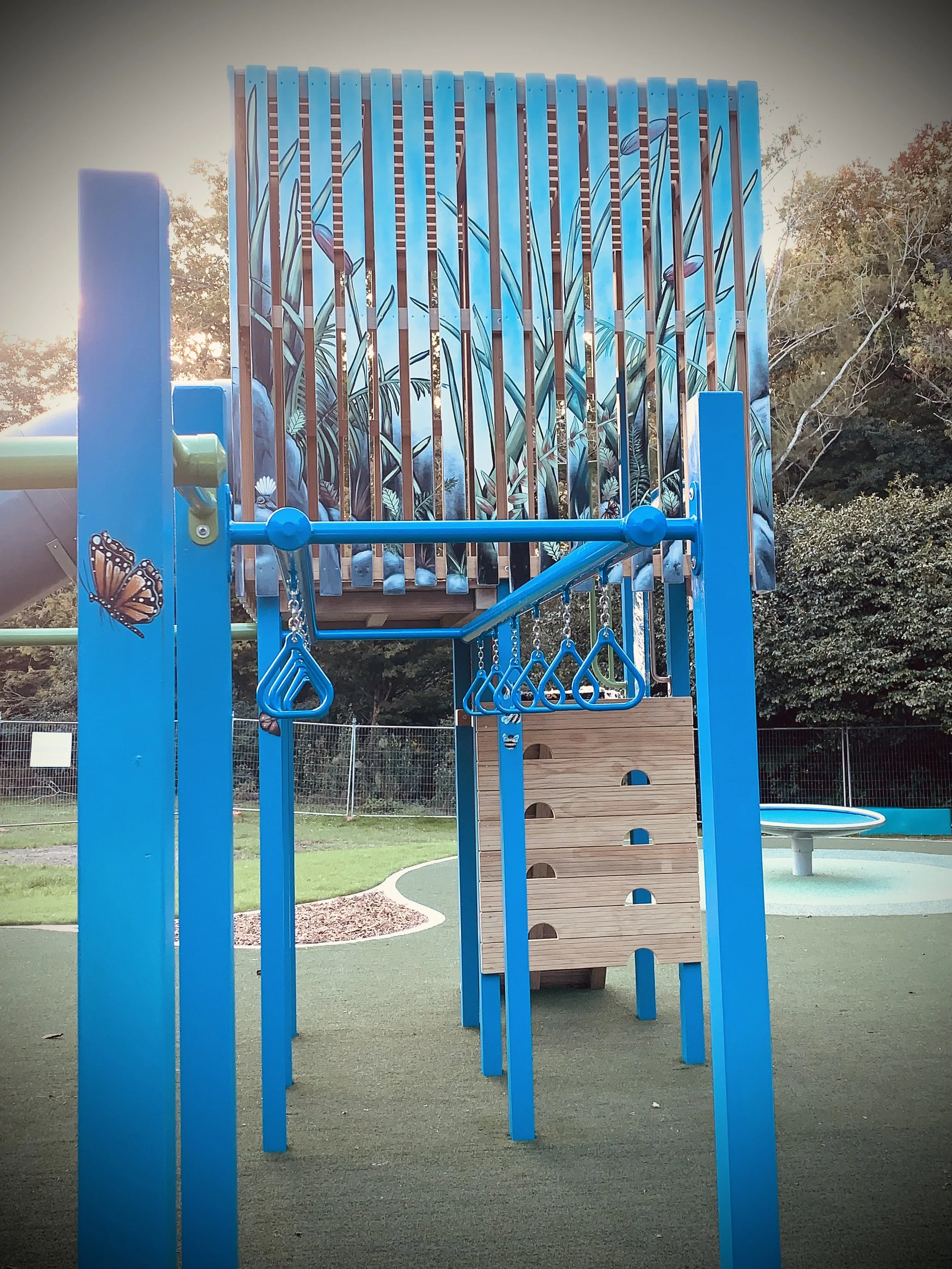 Colorful playground structure with blue and green elements, including monkey bars, a climbing wall, and a slide, set on green rubberized ground surrounded by trees.