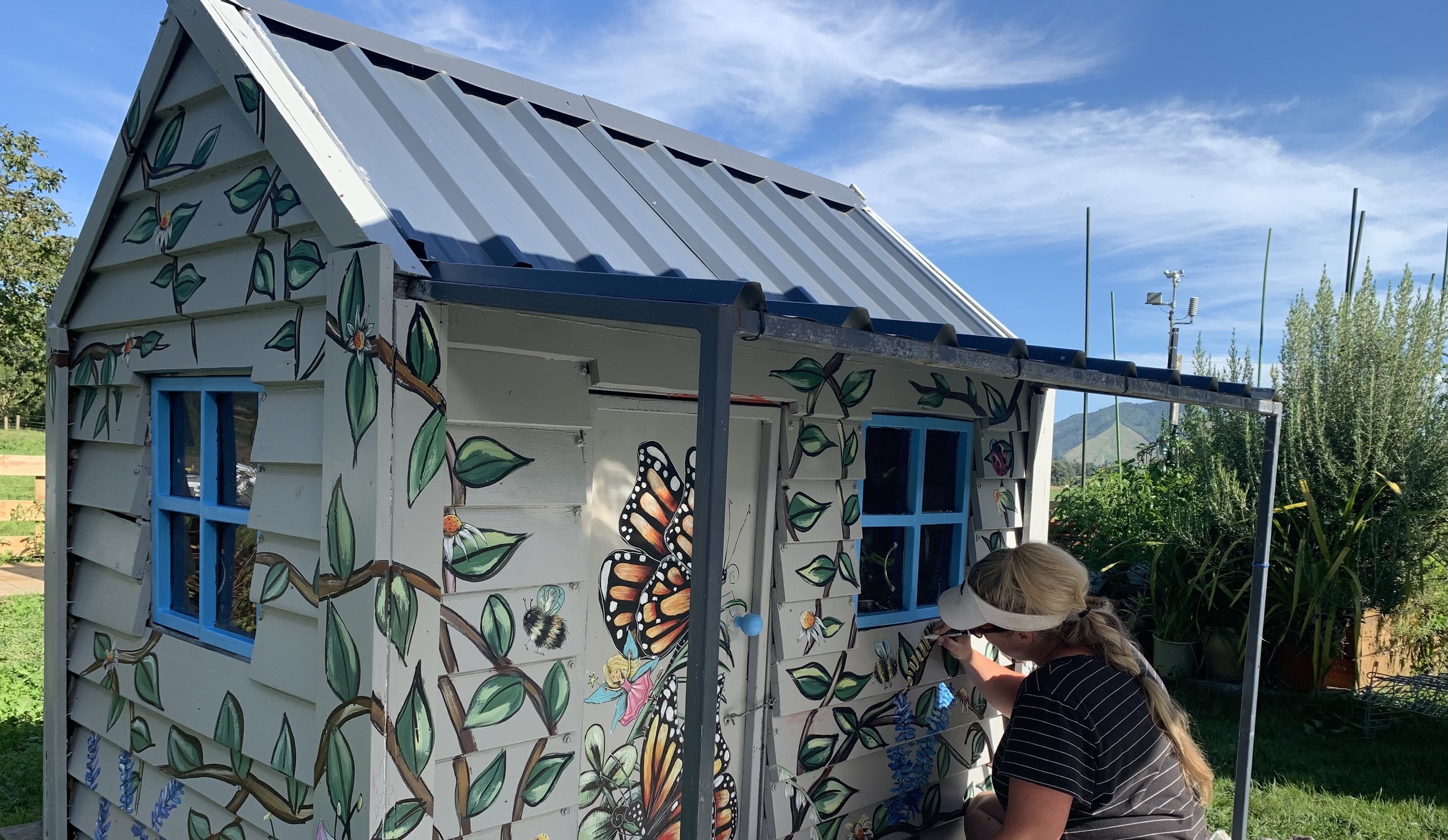 A woman painting butterfly and nature mural on the side of a small garden shed with blue window frames, in a sunny outdoor setting.
