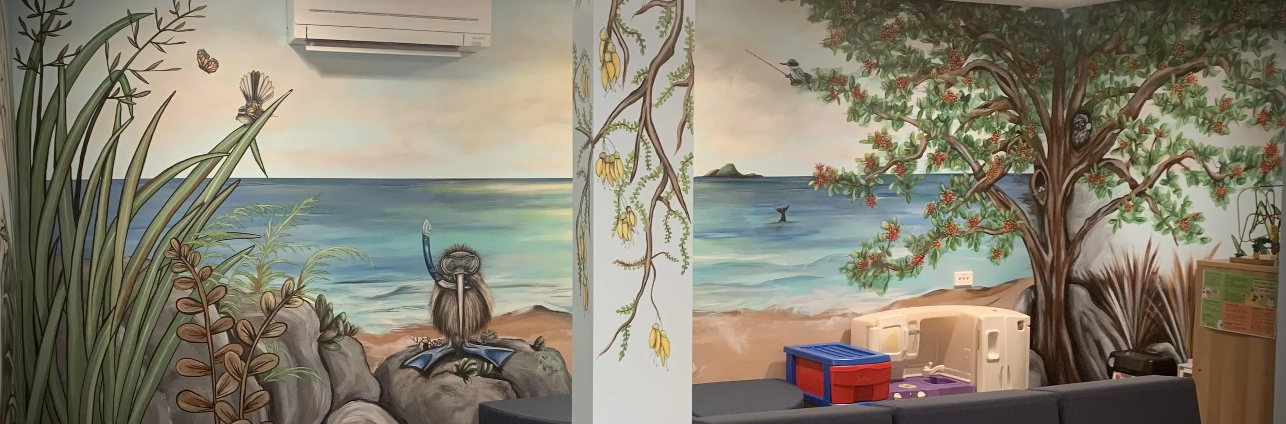 Indoor space with a large mural of a beach scene with water, rocks, and a sky with clouds, painted on the walls. The mural continues across a corner of the room, with the left side showing tall grasses, a butterfly, and a person with a snorkel sittin