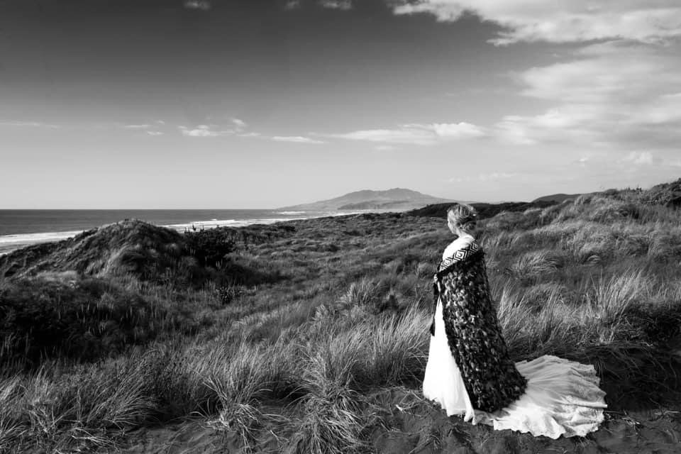 Dannika on her wedding day, standing on a grassy coastal landscape near the ocean, wearing a cloak, looking into the distance.