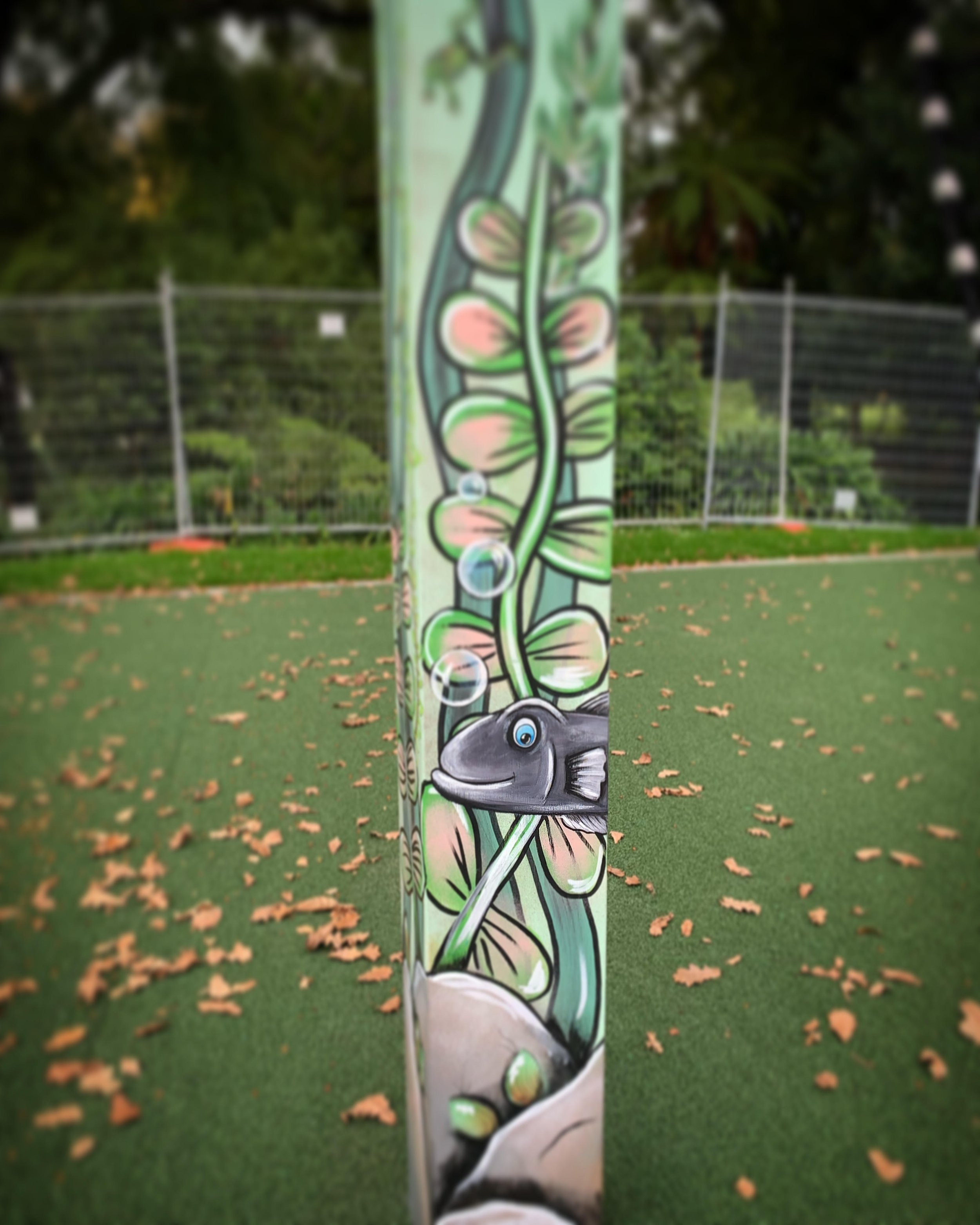Colorful mural of a fish with bubbles, plants, and rocks on a vertical surface in a park with a grassy area and a fence in the background.