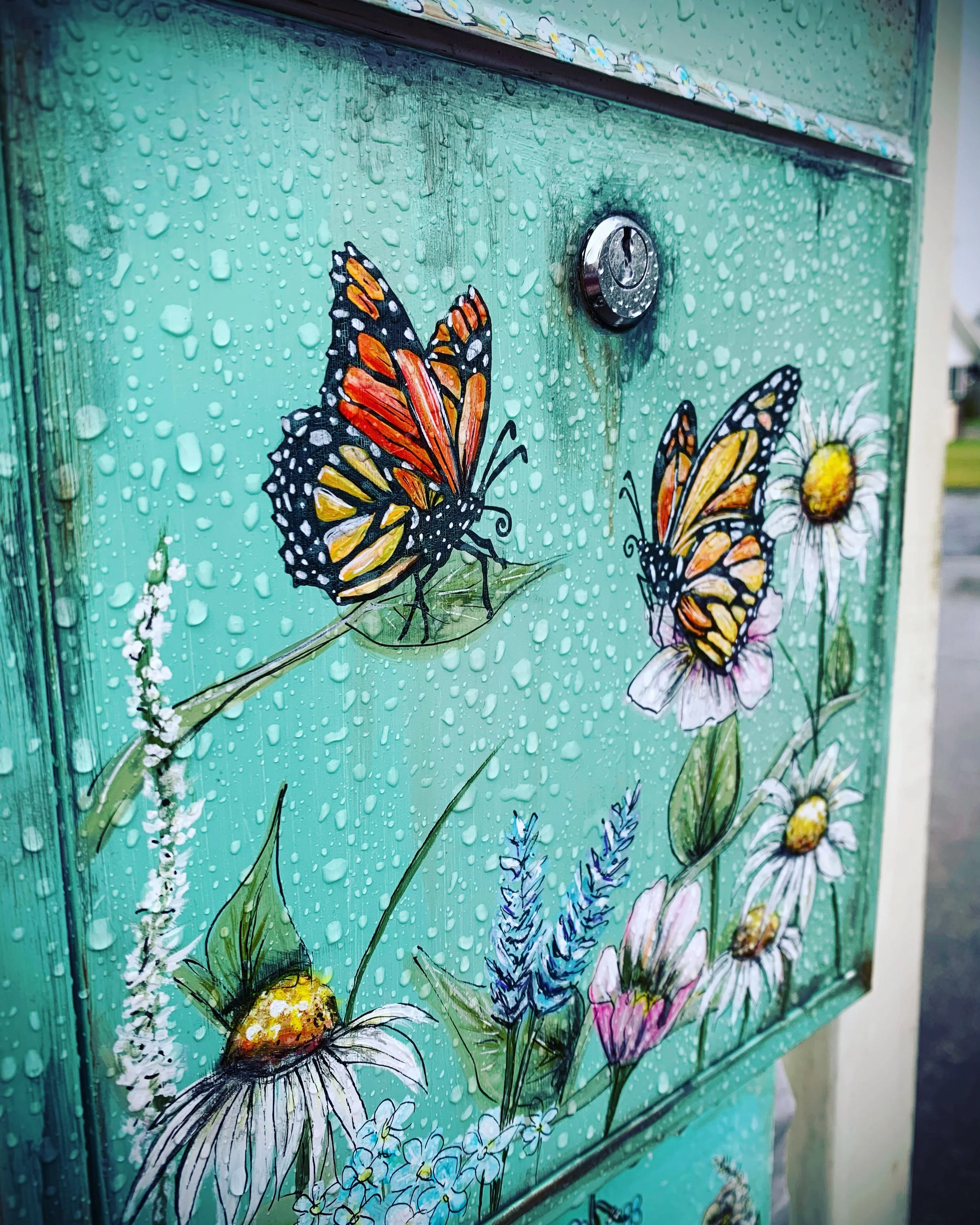 Colorful mailbox with rain droplets, decorated with painted orange and black monarch butterflies and white daisies and other wildflowers.