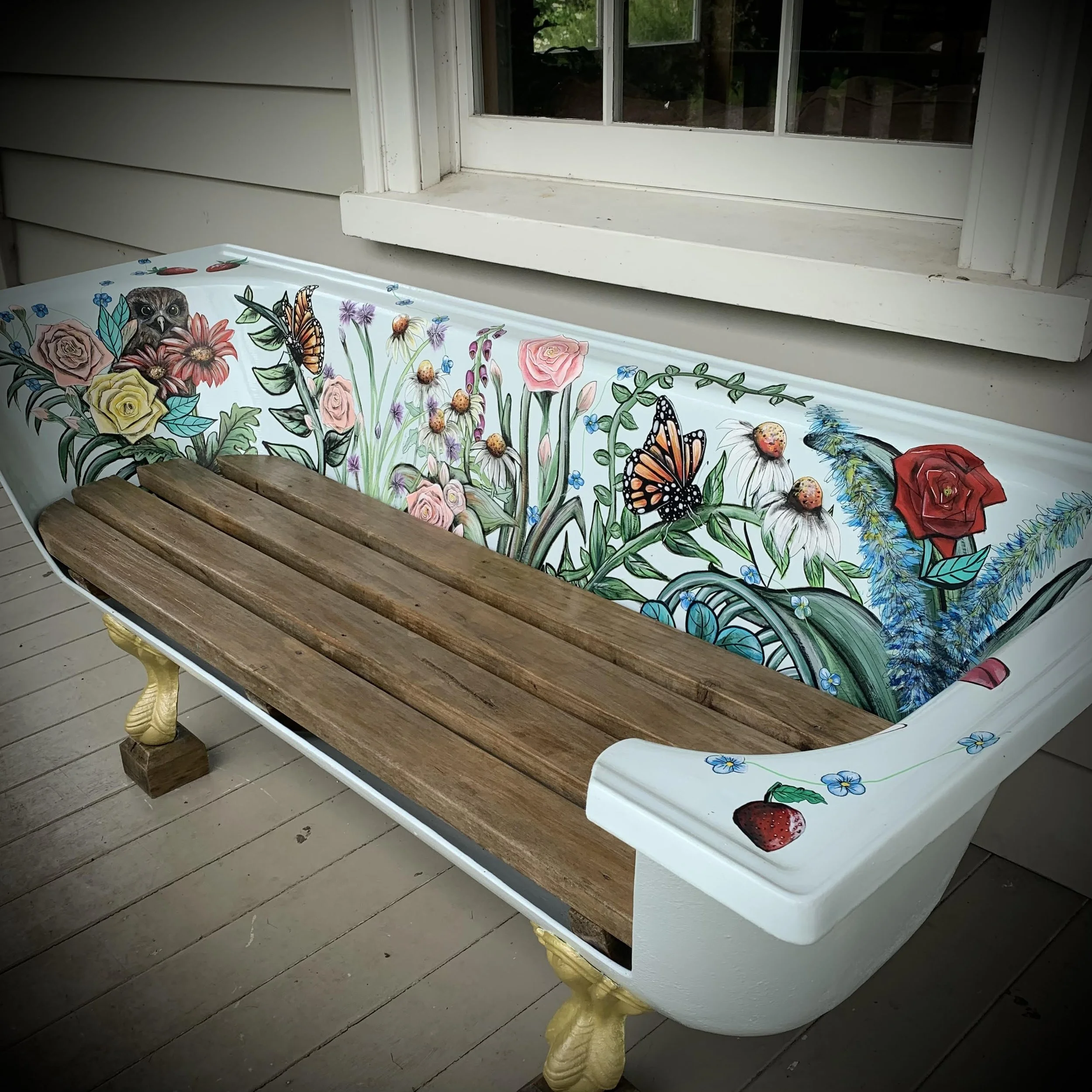 A porch swing with a colorful painted backrest featuring flowers, butterflies, an owl, and a strawberry, with a wooden slat seat and decorative yellow legs.