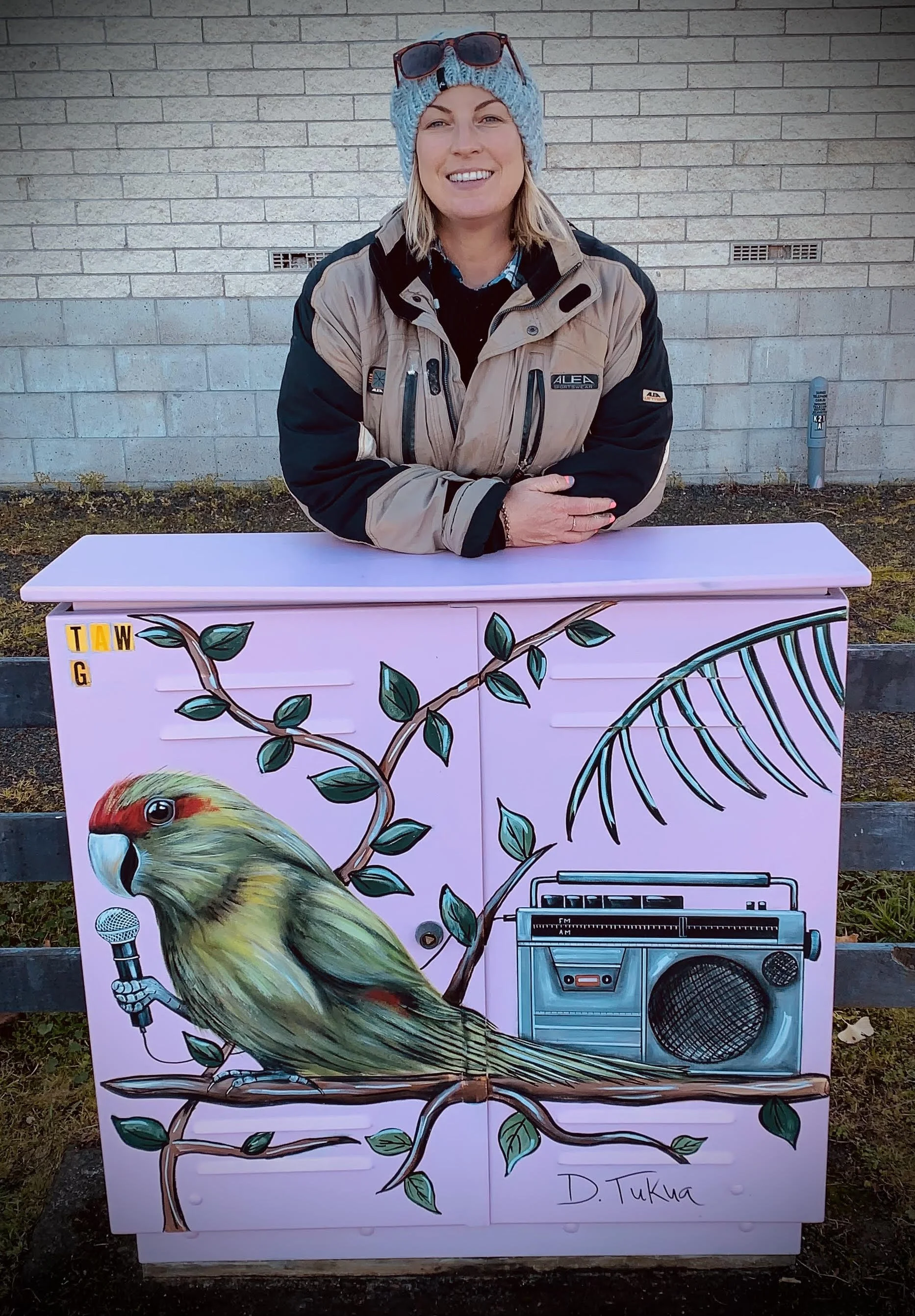 A woman wearing a blue knit hat, sunglasses, and a beige and black jacket stands behind a painted pink utility box with a bird holding a microphone, a tree branch, leaves, a fern, and a boombox, signed D. Tukua.
