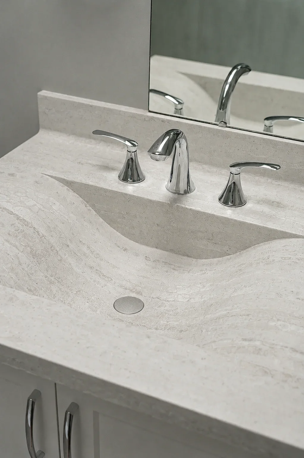 A bathroom sink with a polished chrome faucet and handles on a light beige stone countertop, reflected in a mirror.